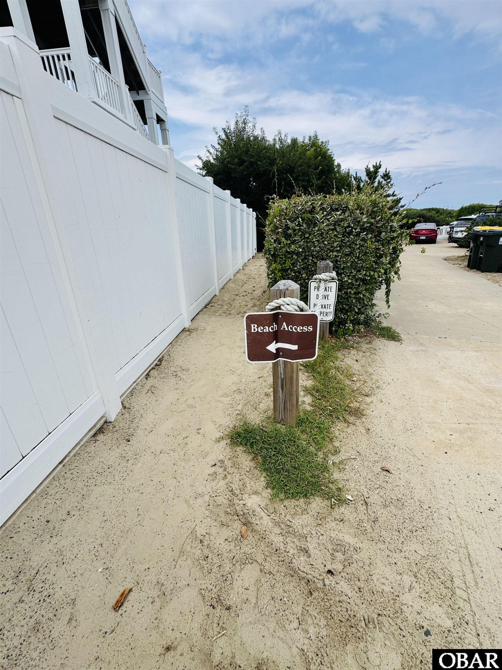 Beach access sign points left beside a sandy path and white fence.