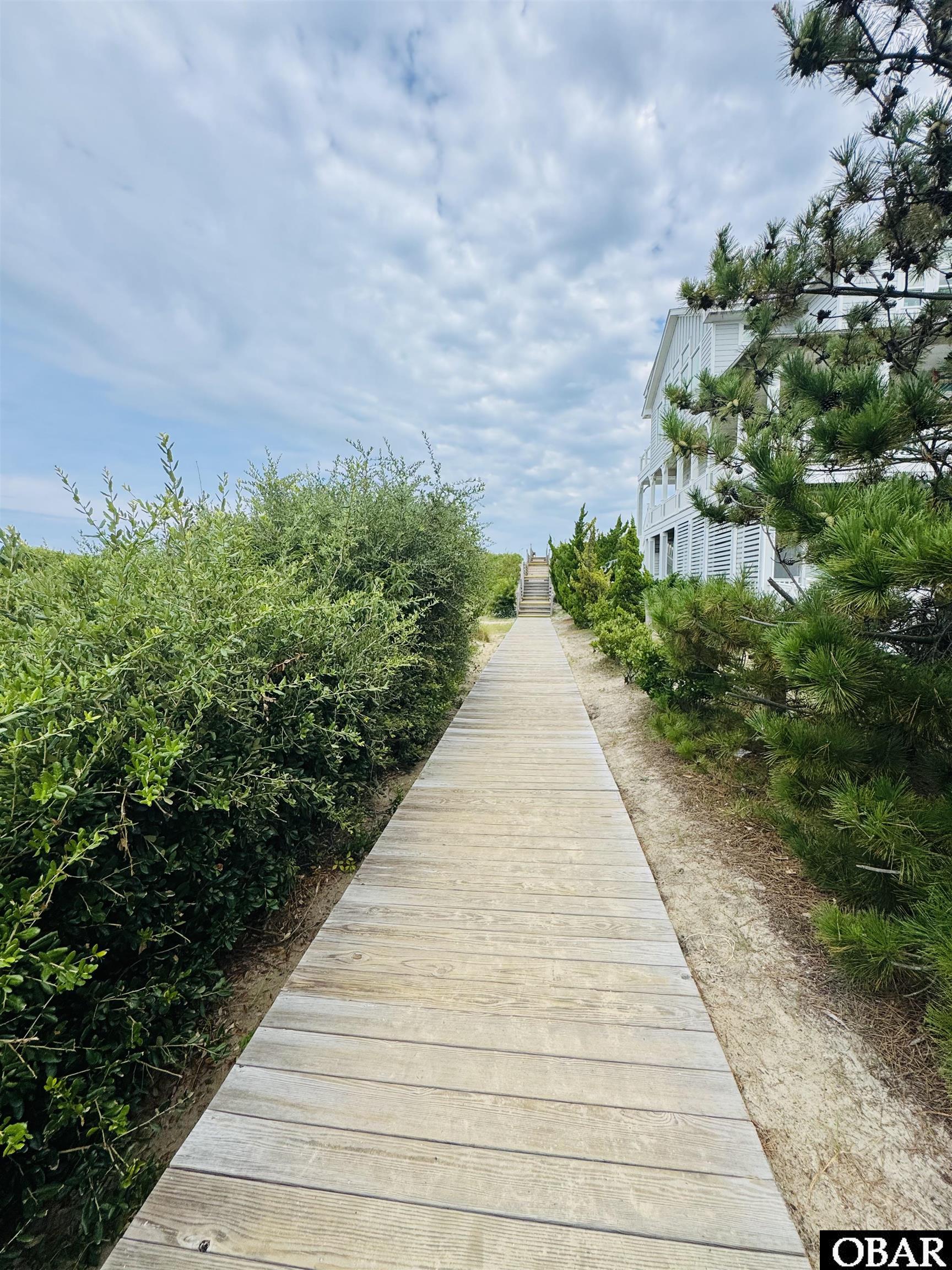 White multi-level house beside a wooden boardwalk bordered by shrubs and pine trees.