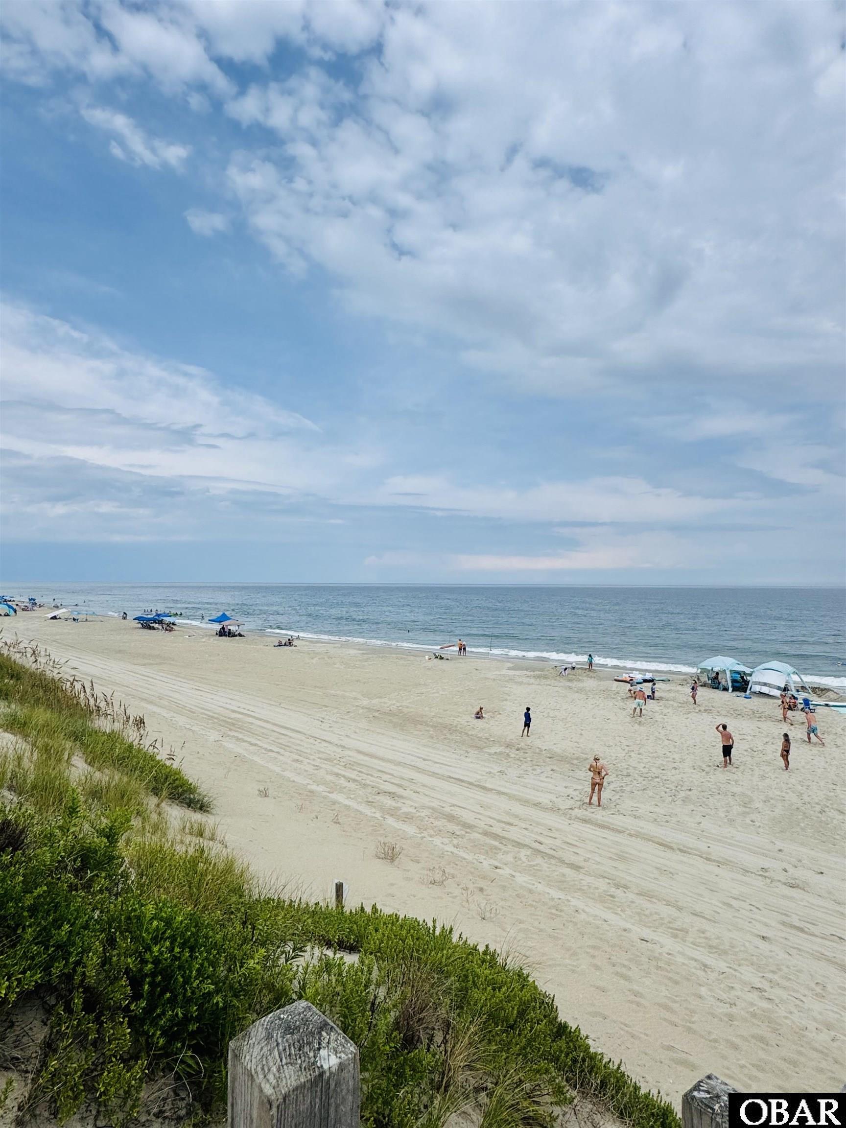 Sandy beach and ocean viewed from grassy dunes, with beachgoers and canopies along the shore.