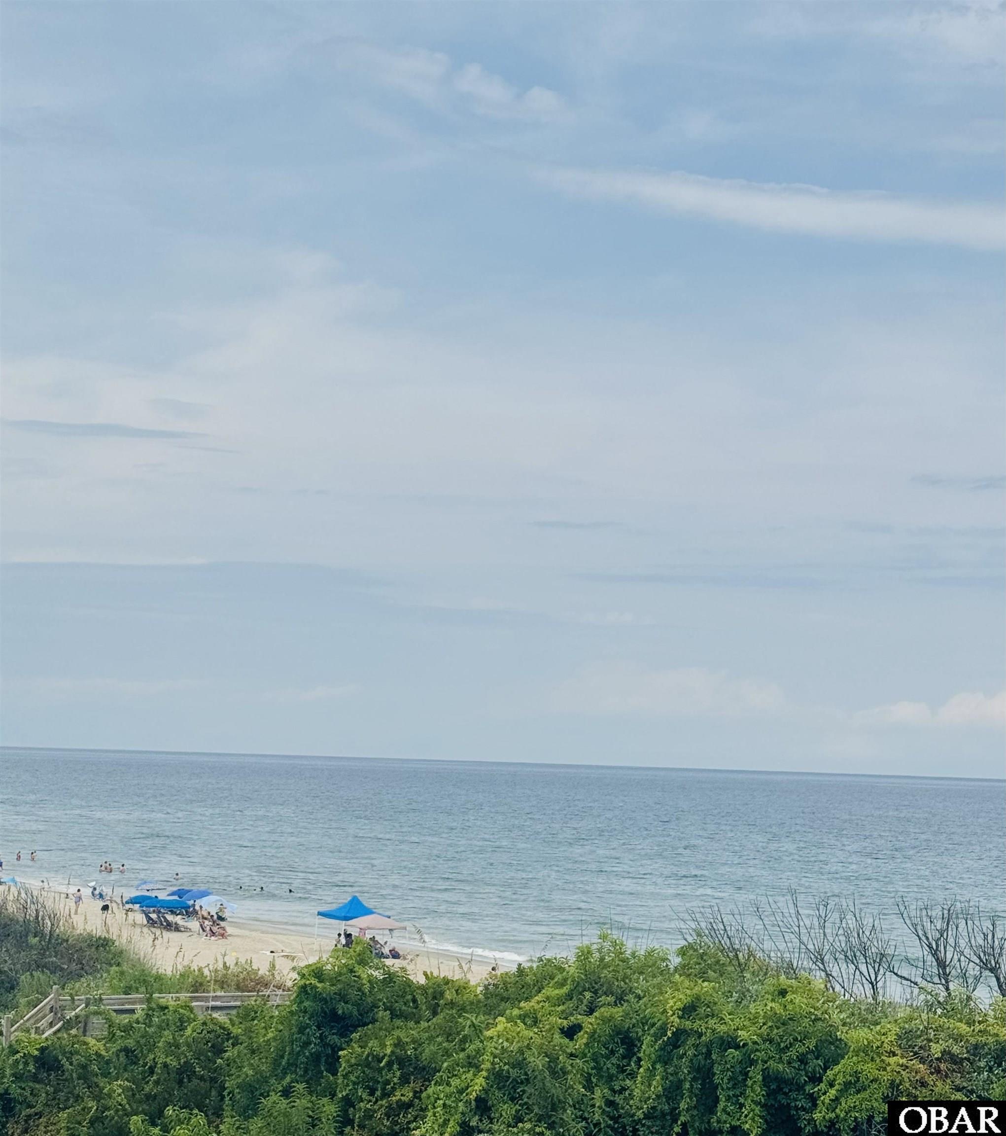 Ocean and beach view with colorful umbrellas, swimmers, and dense dune greenery.