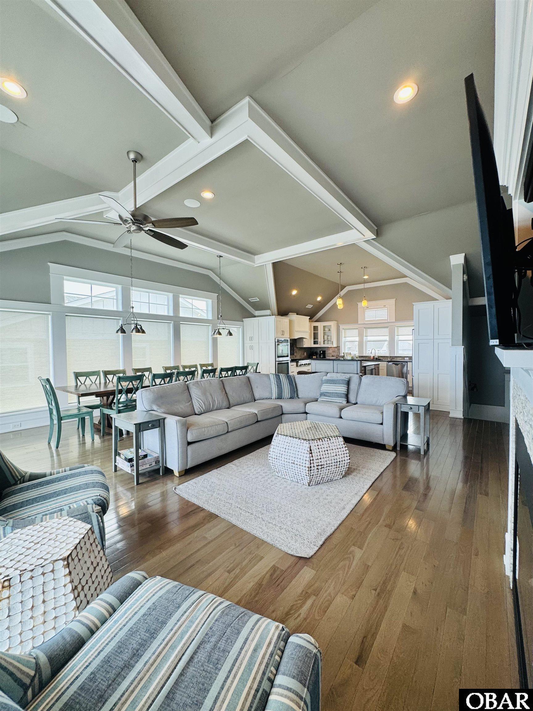 Open living room with gray sectional, hardwood floors, vaulted ceiling, and dining area beside the kitchen.