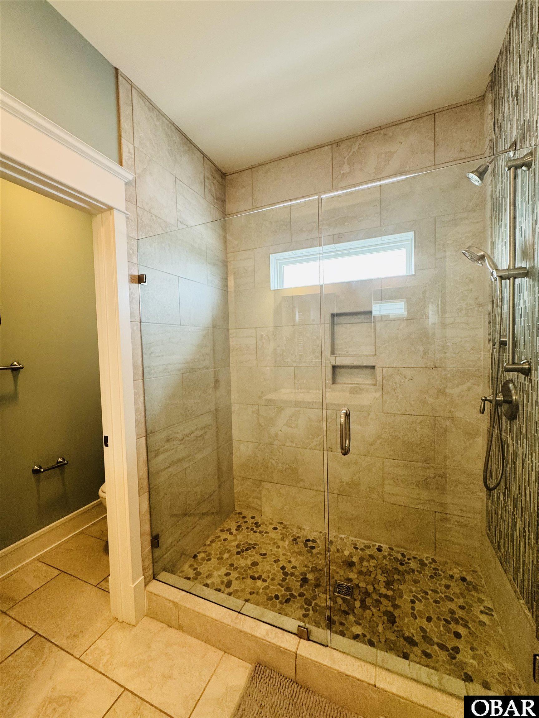 Walk-in shower with glass doors, beige stone tile, pebble floor, built-in niches, and mosaic accent wall.