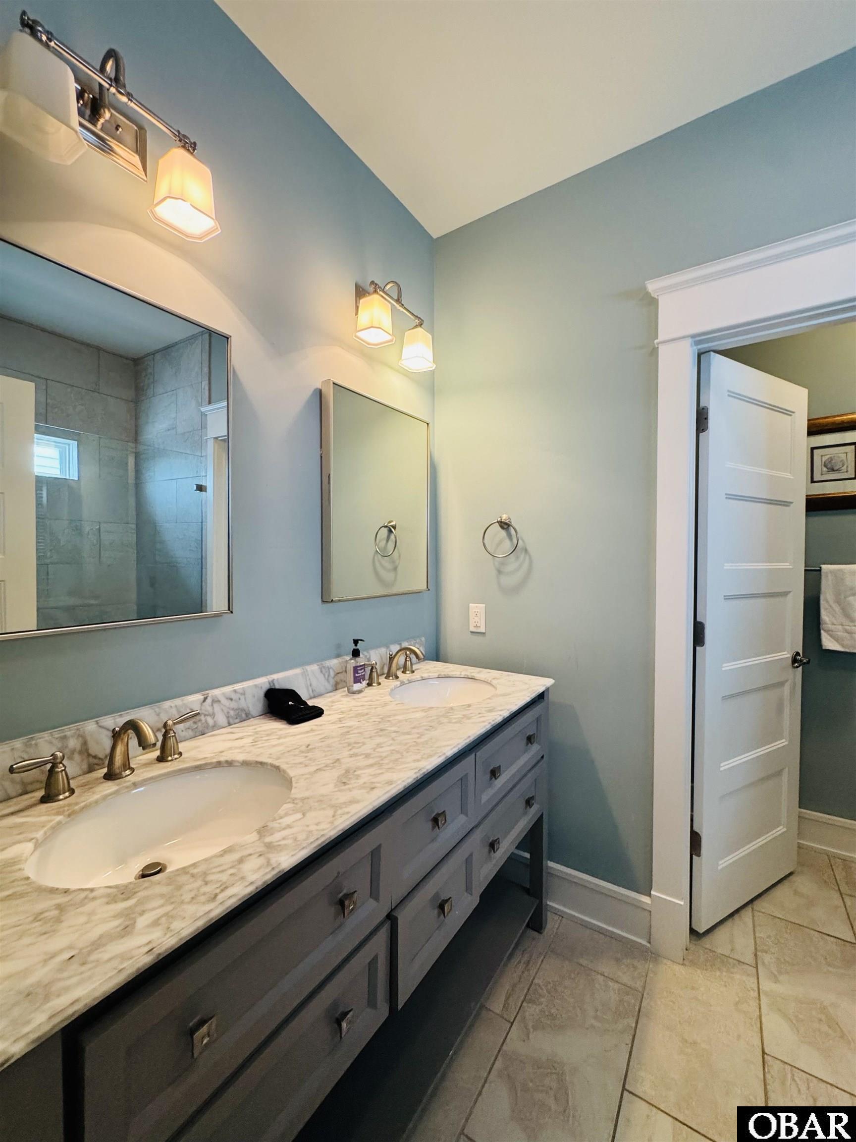 Bathroom with a double-sink marble vanity, gray cabinets, blue-green walls, and a tiled shower reflected in the mirror.