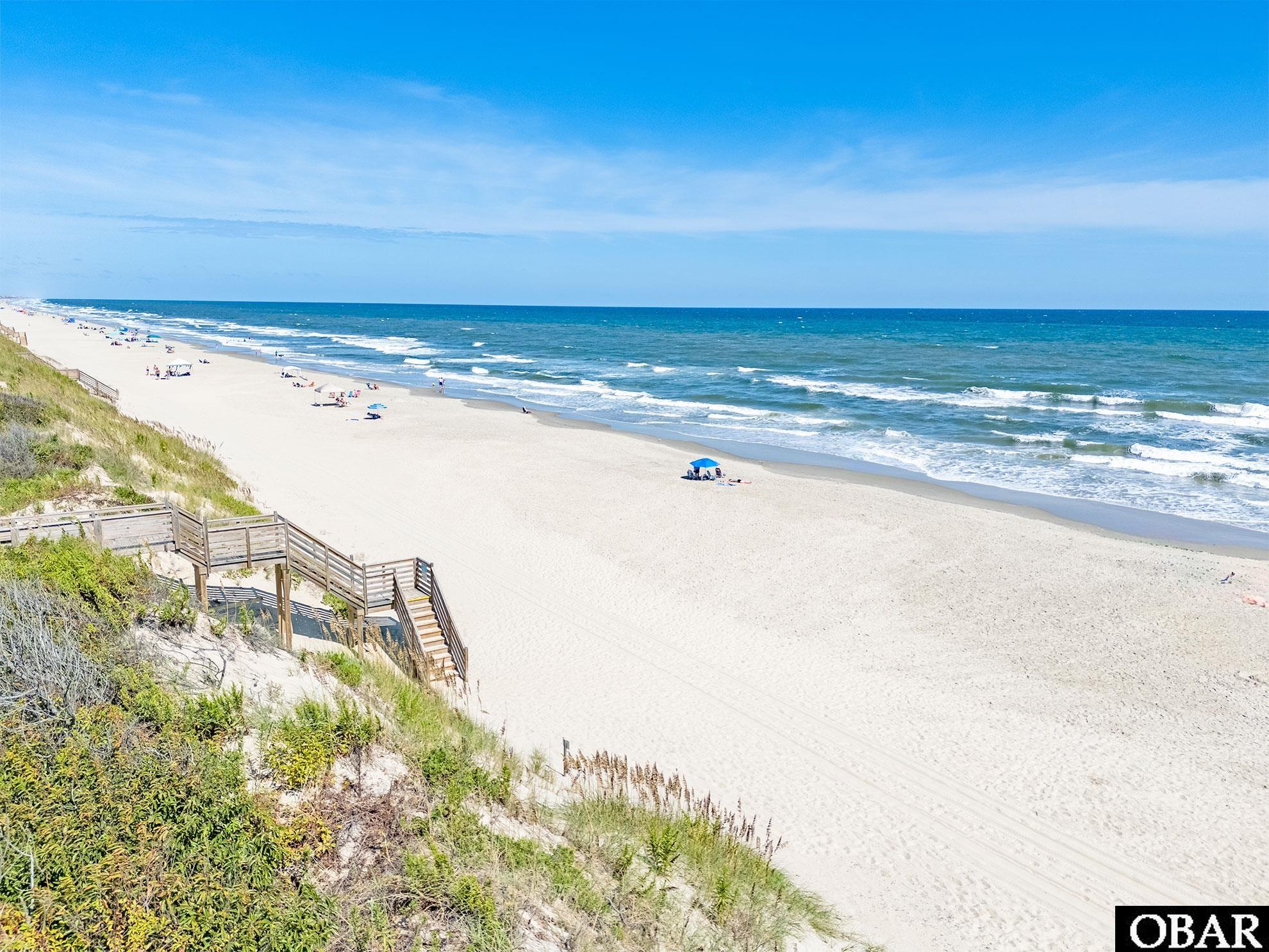 Sandy beach and ocean with a wooden dune staircase and scattered beach umbrellas.