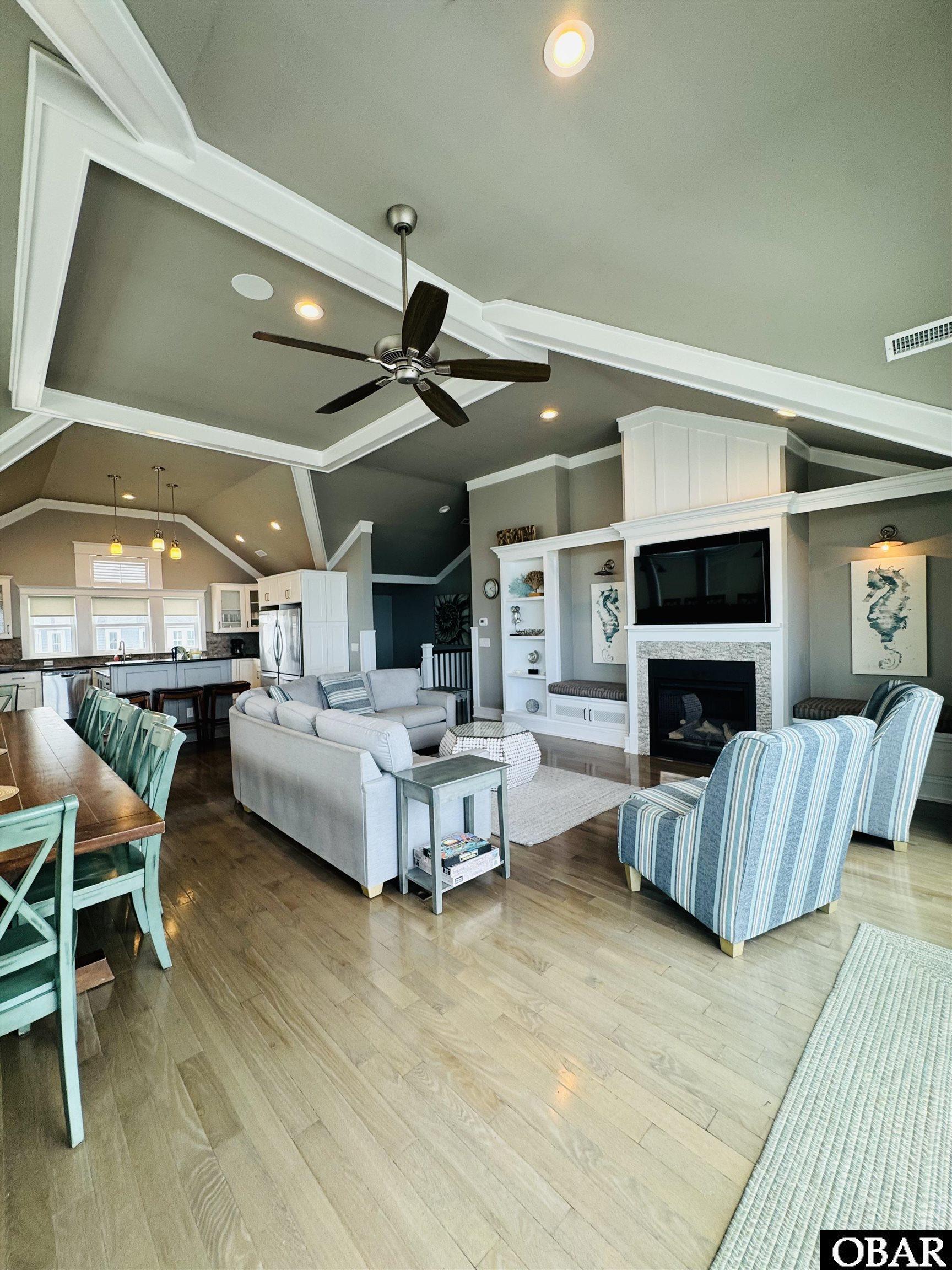 Open-plan living room with a stone fireplace, built-ins, blue-striped armchairs and kitchen dining area.