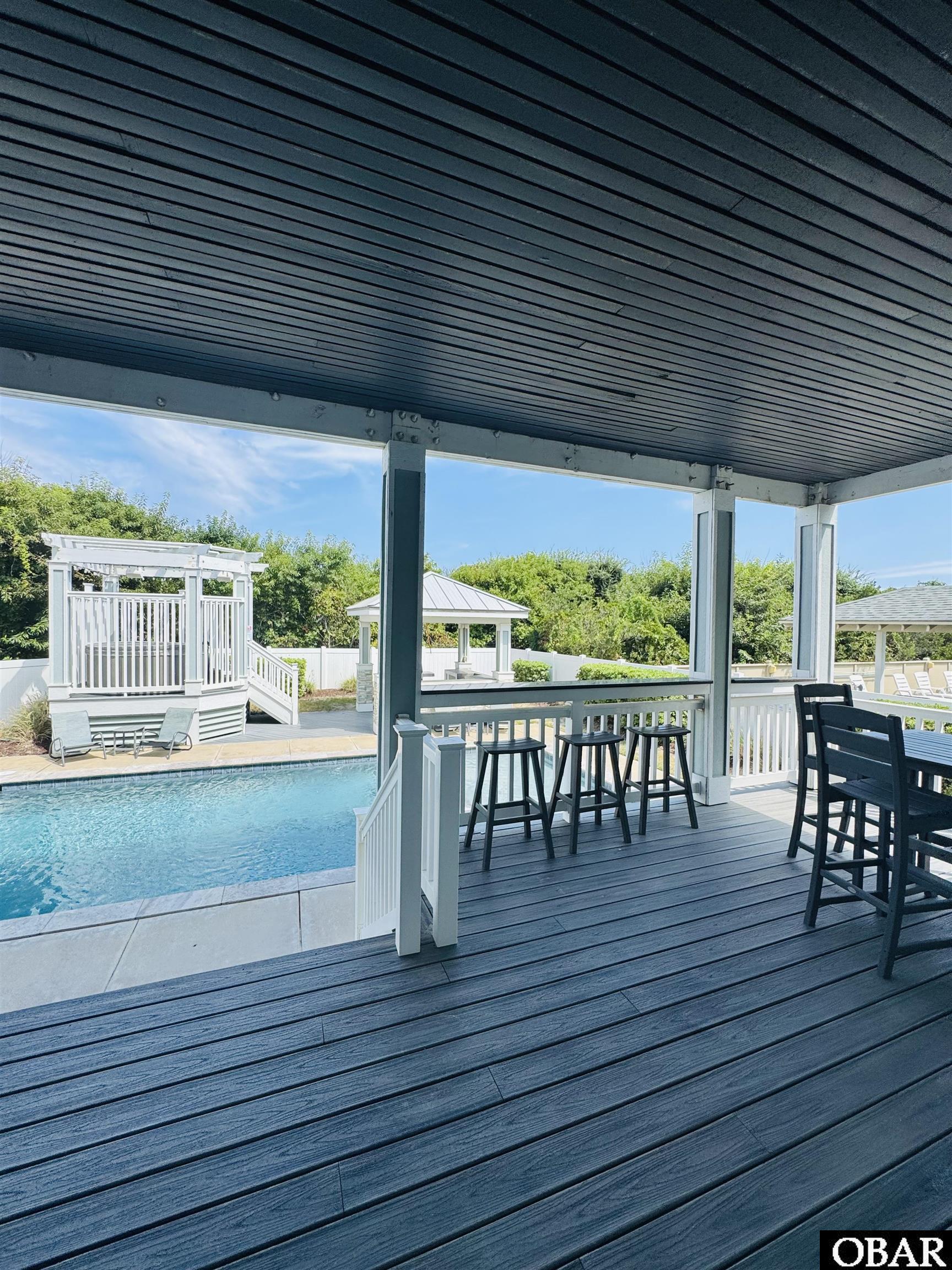 Covered deck with black bar stools overlooking a swimming pool and white gazebo.