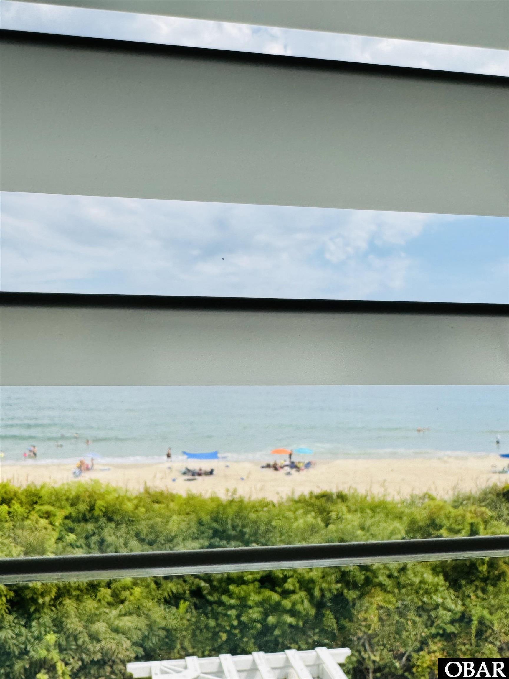 Ocean and beach view framed by porch railings, with dune vegetation and beachgoers below.