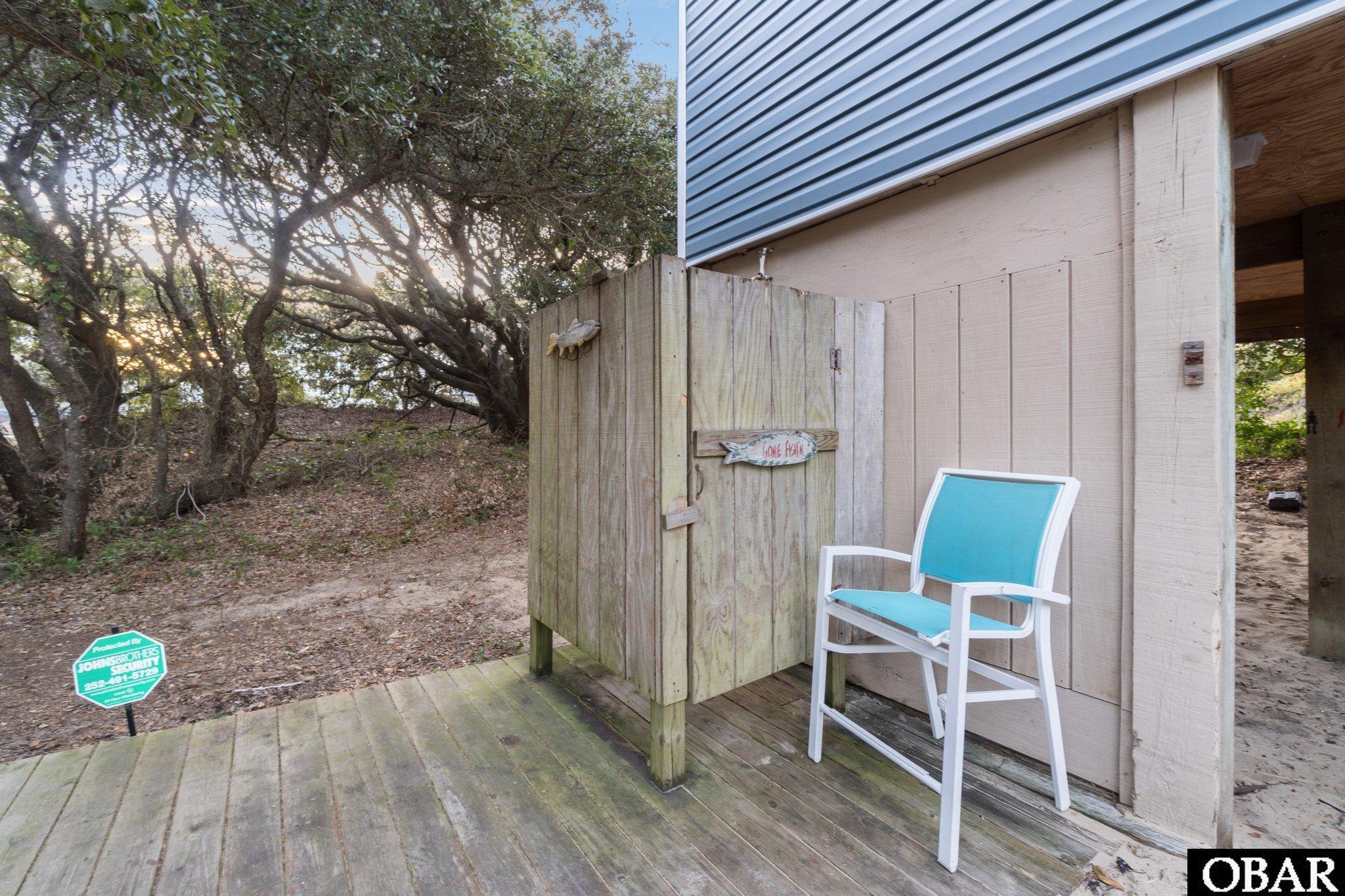 Wooden deck with a screened outdoor shower and turquoise chair beside mature trees.