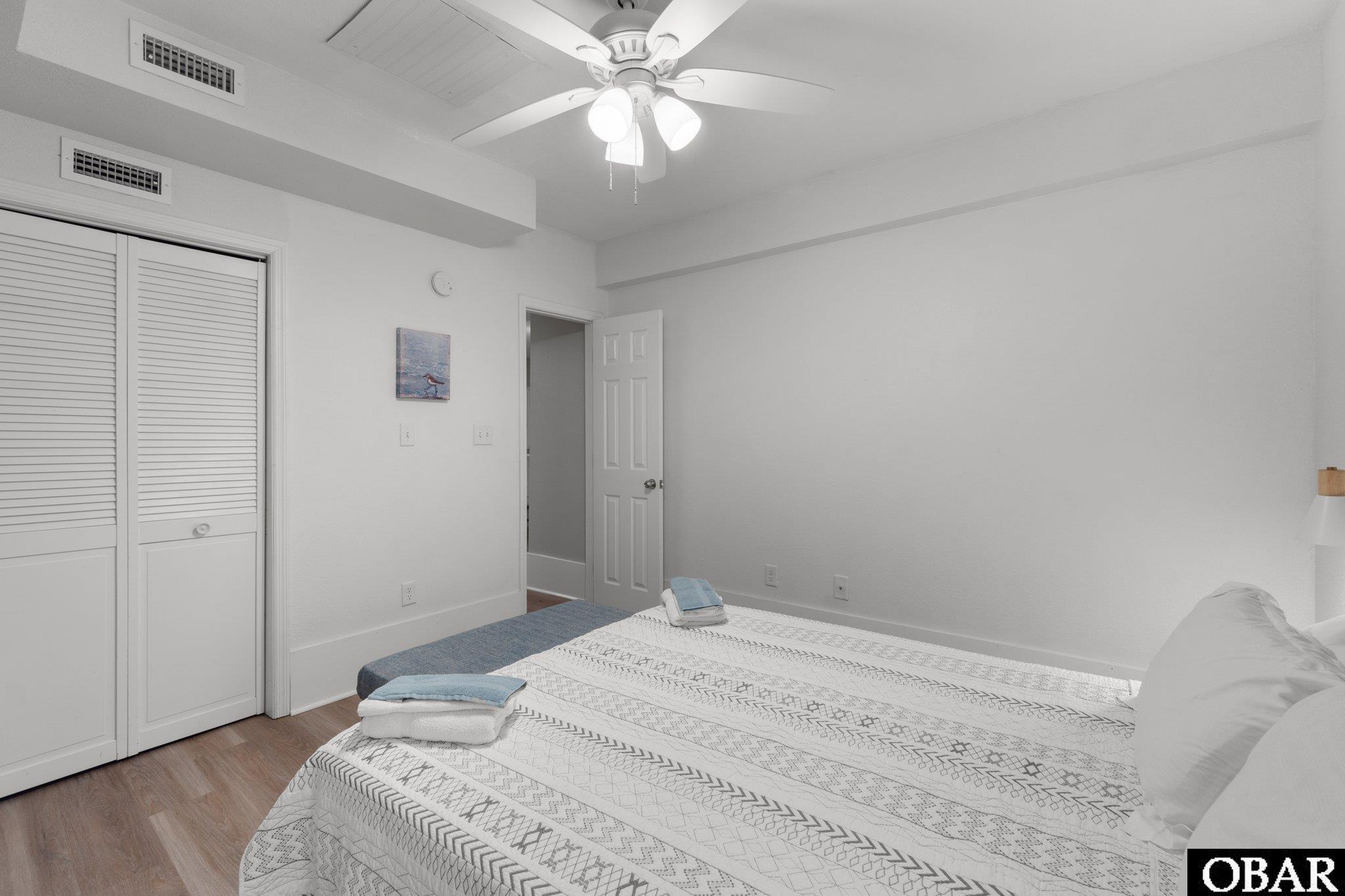 Bedroom with a patterned white quilt, folded towels, and louvered closet doors.