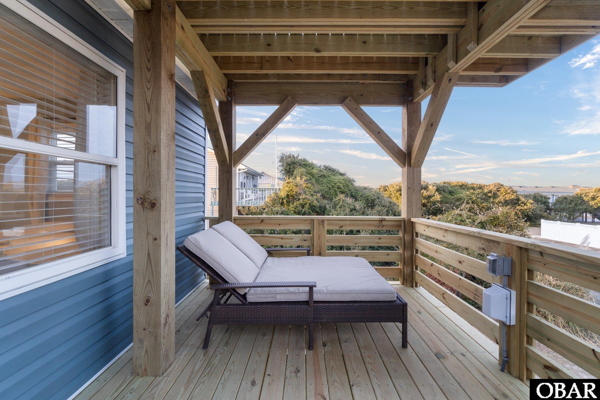 Covered wooden deck with a cushioned chaise lounge and views of shrubs and neighboring homes.