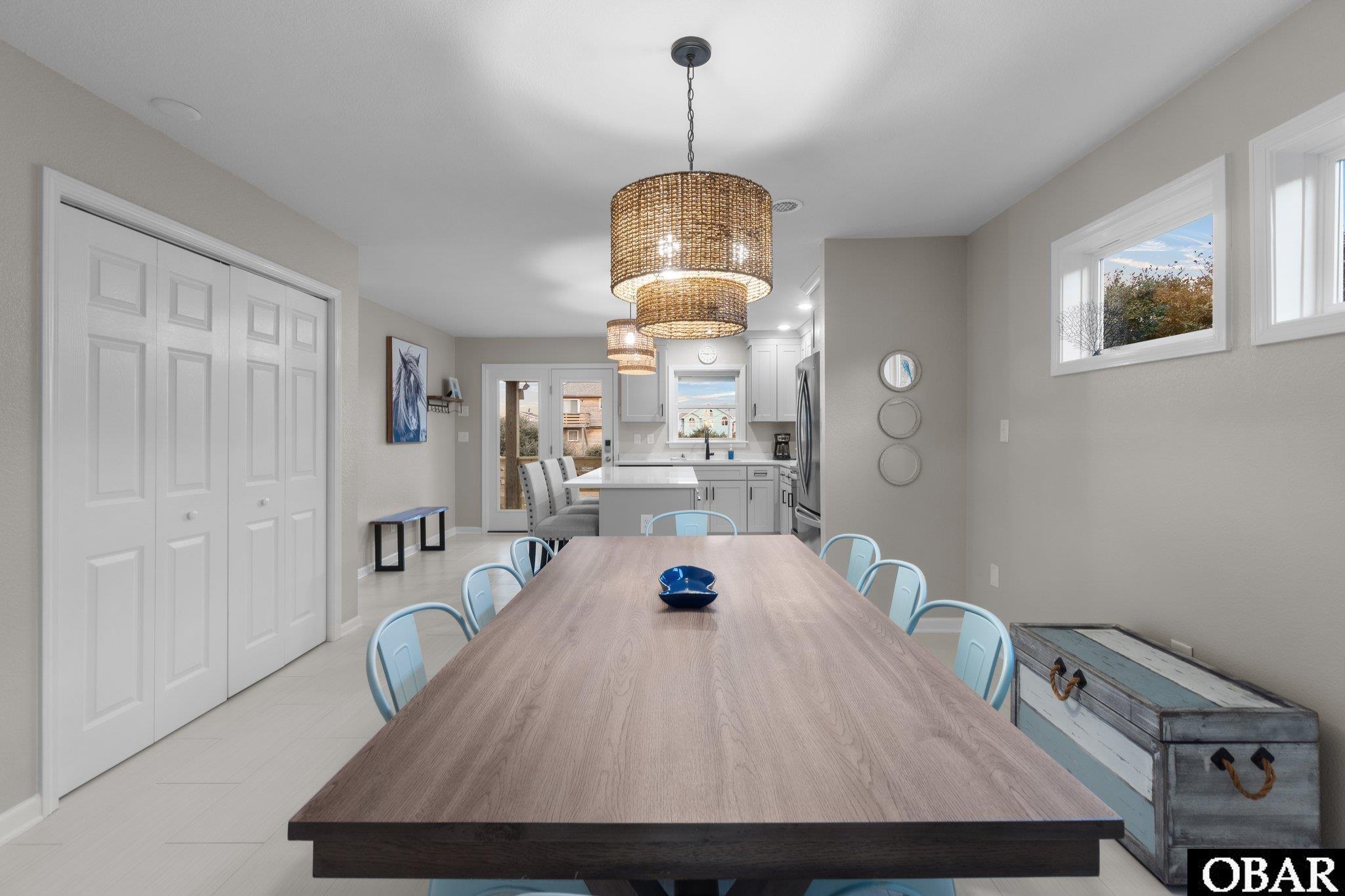 Dining room with a long wood table, pale blue chairs, woven pendant lights, and an adjoining white kitchen.
