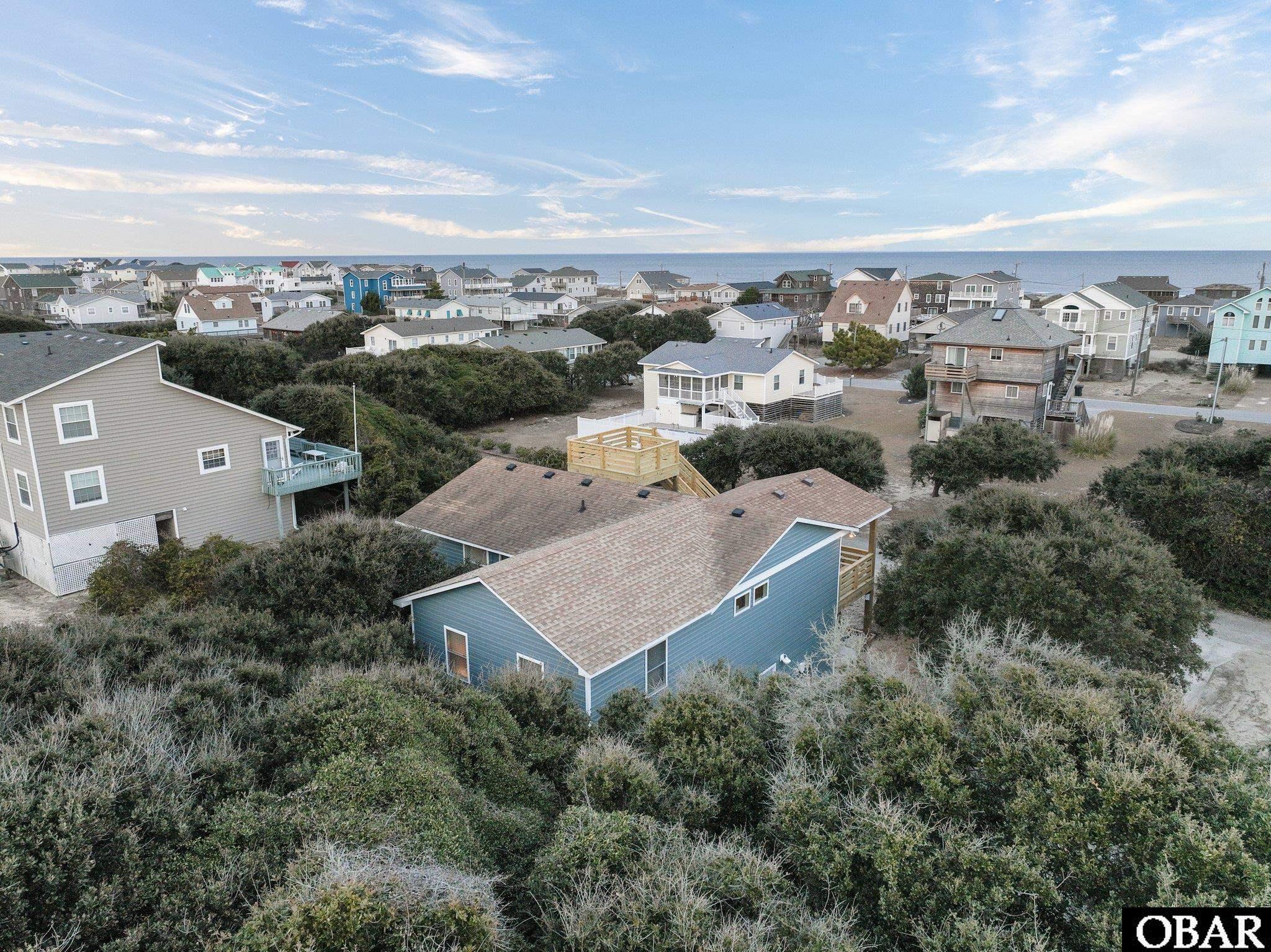 Aerial view of a blue coastal house among dune shrubs, with neighboring homes and ocean beyond.