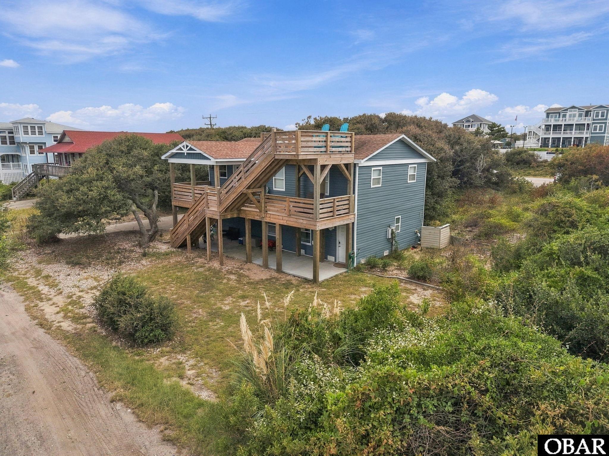 Blue coastal house with broad wooden decks, exterior stairs, and rooftop seating amid dune vegetation.