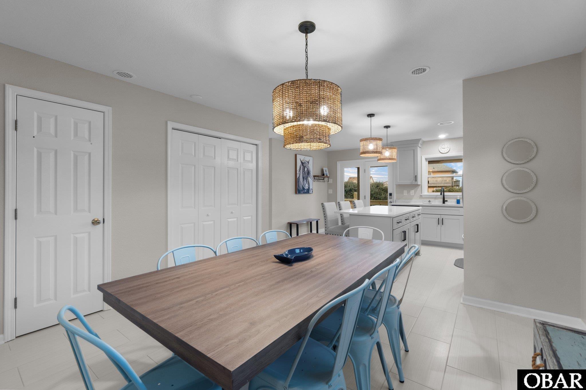 Dining area with a long wood table, blue chairs, woven pendant lights, and an adjoining white kitchen.