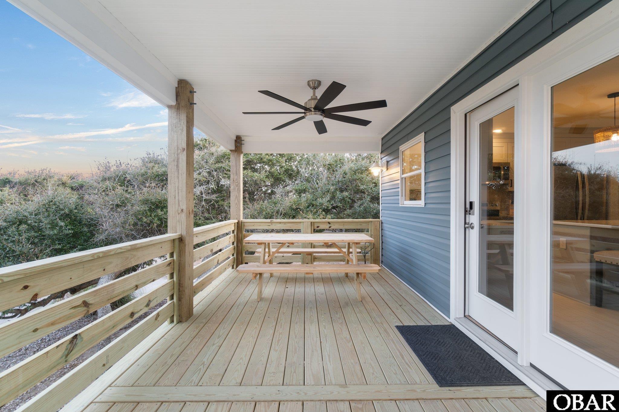 Covered wooden porch with a picnic table, ceiling fan, and blue siding beside glass doors.