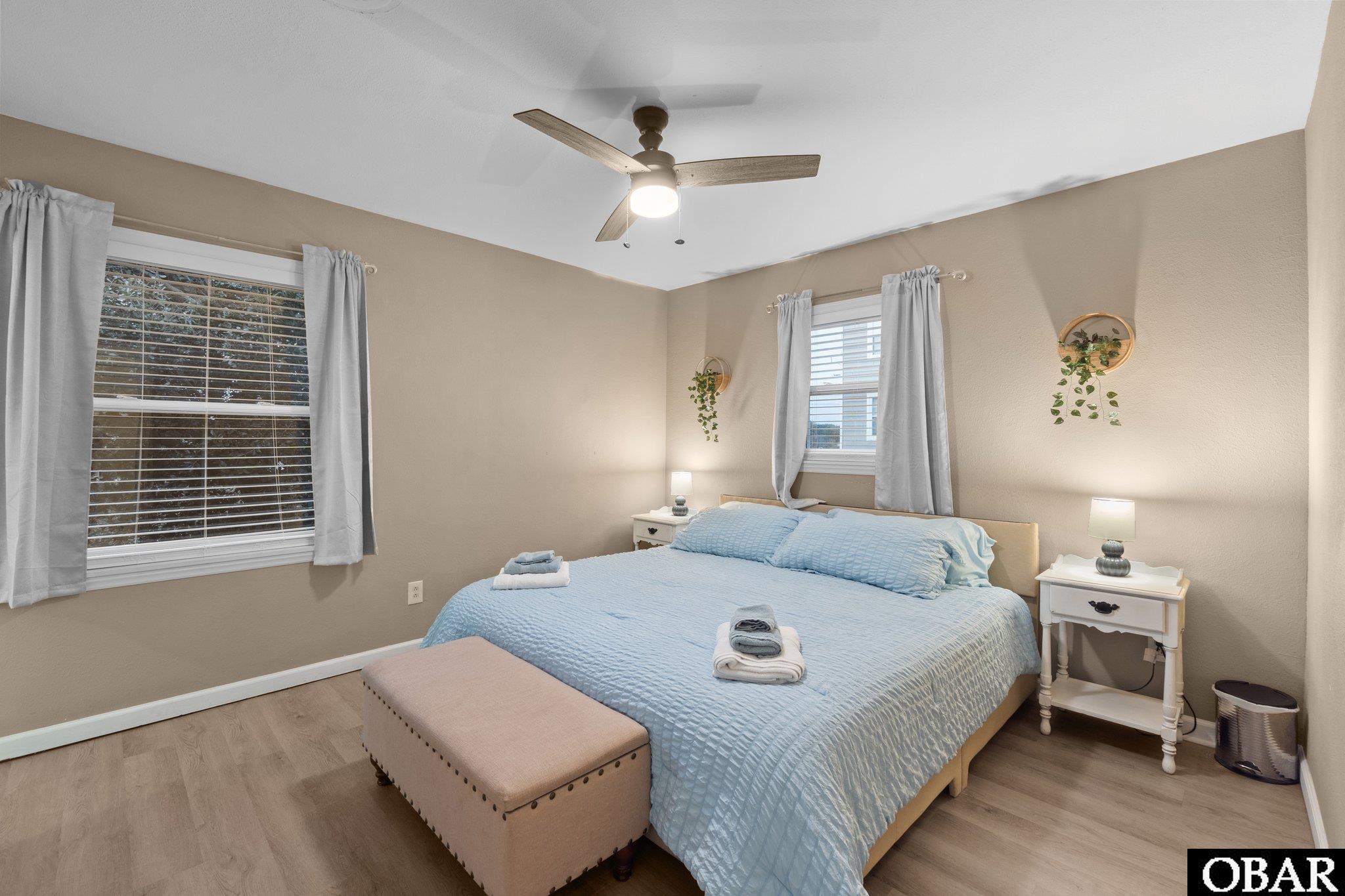 Bedroom with a pale blue bed, upholstered bench, curtained windows, and ceiling fan.