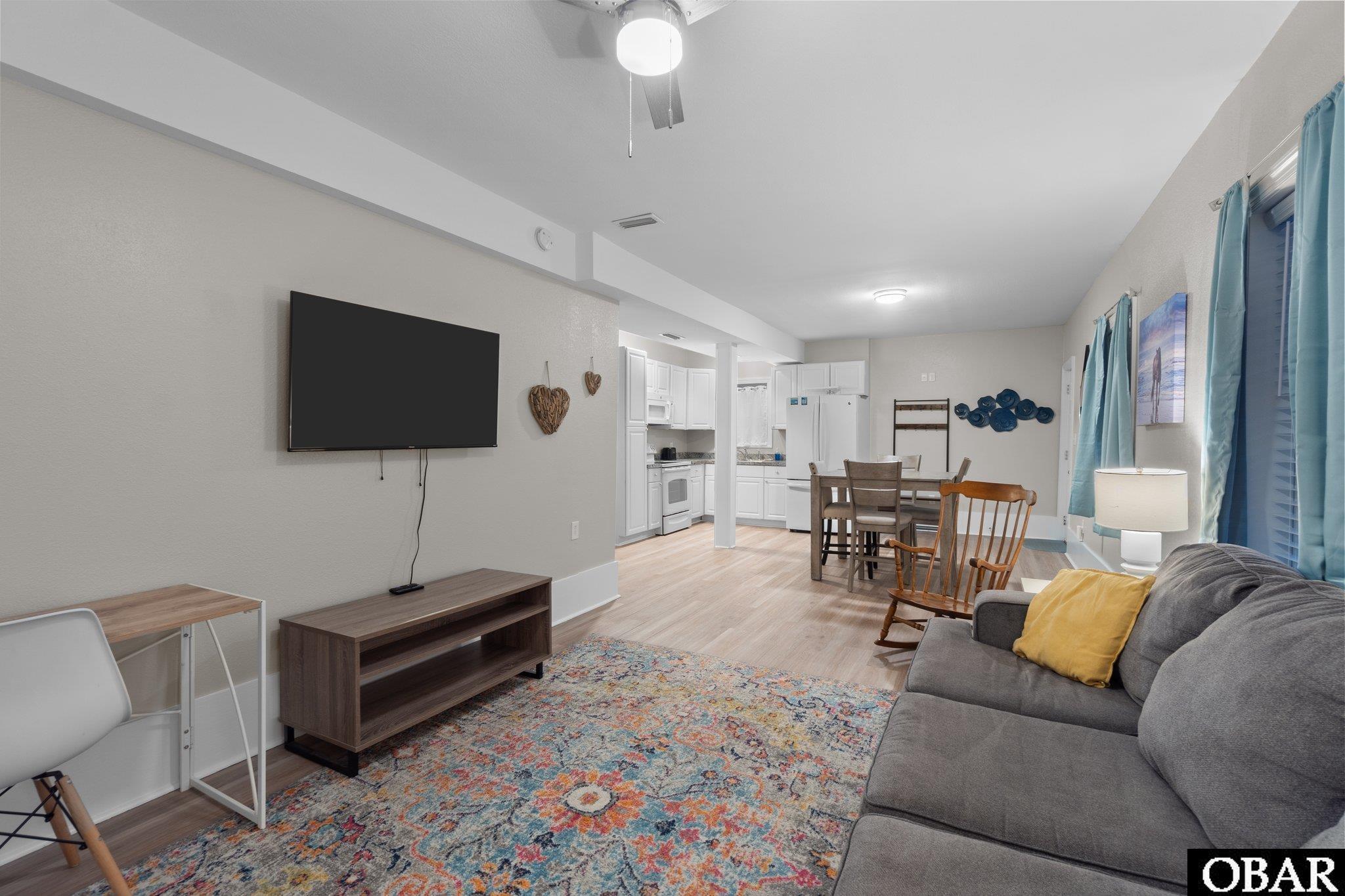 Open-plan living and dining area with a gray sectional sofa, colorful rug, and white kitchen.