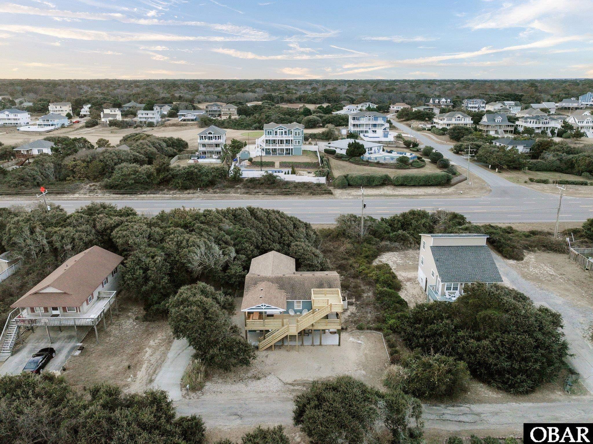 Aerial view of a coastal home with a large raised deck, scrub vegetation, and neighboring houses across the road.