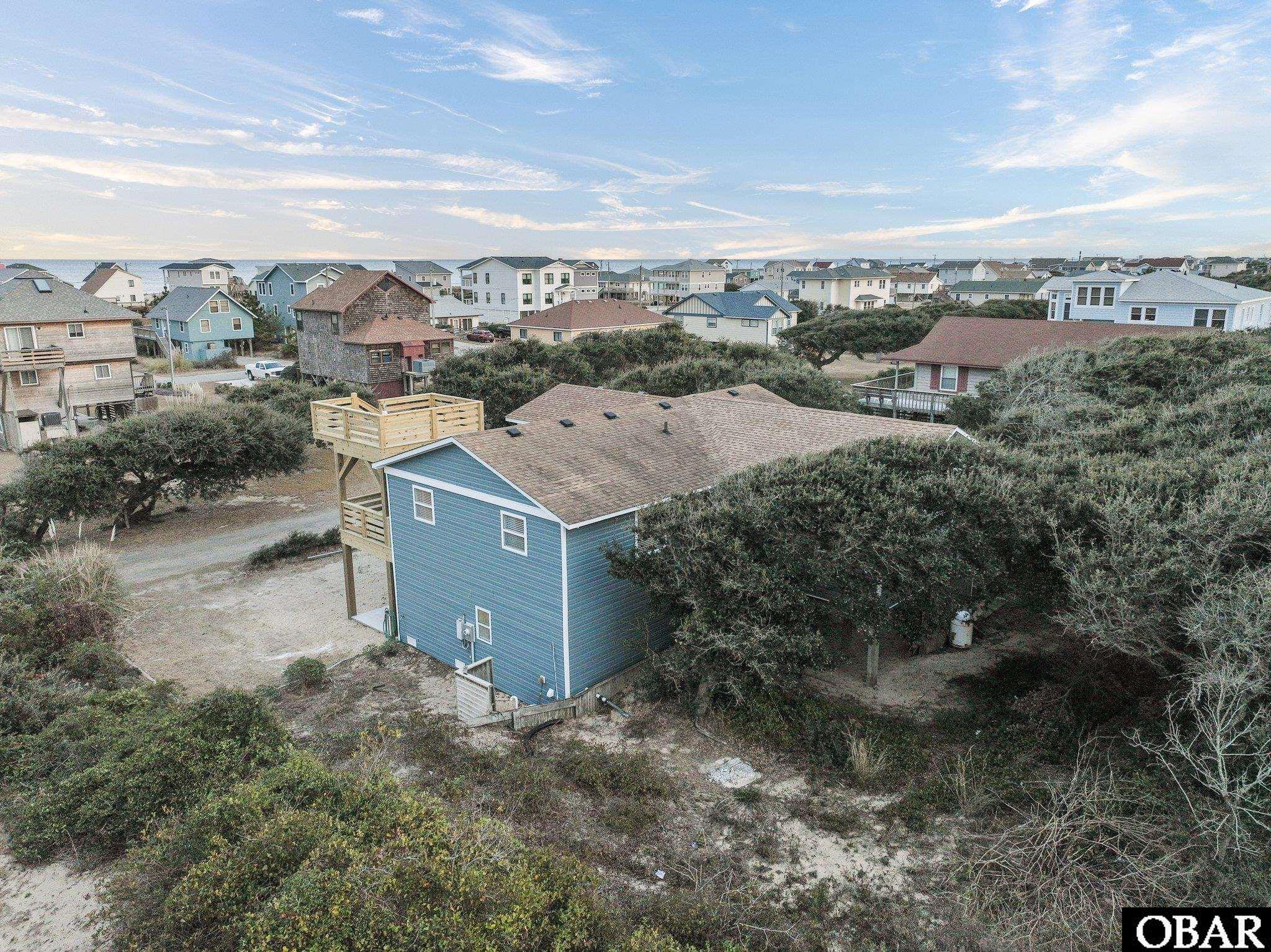 Blue coastal house with a raised rooftop deck amid dune vegetation, neighboring homes, and ocean views.