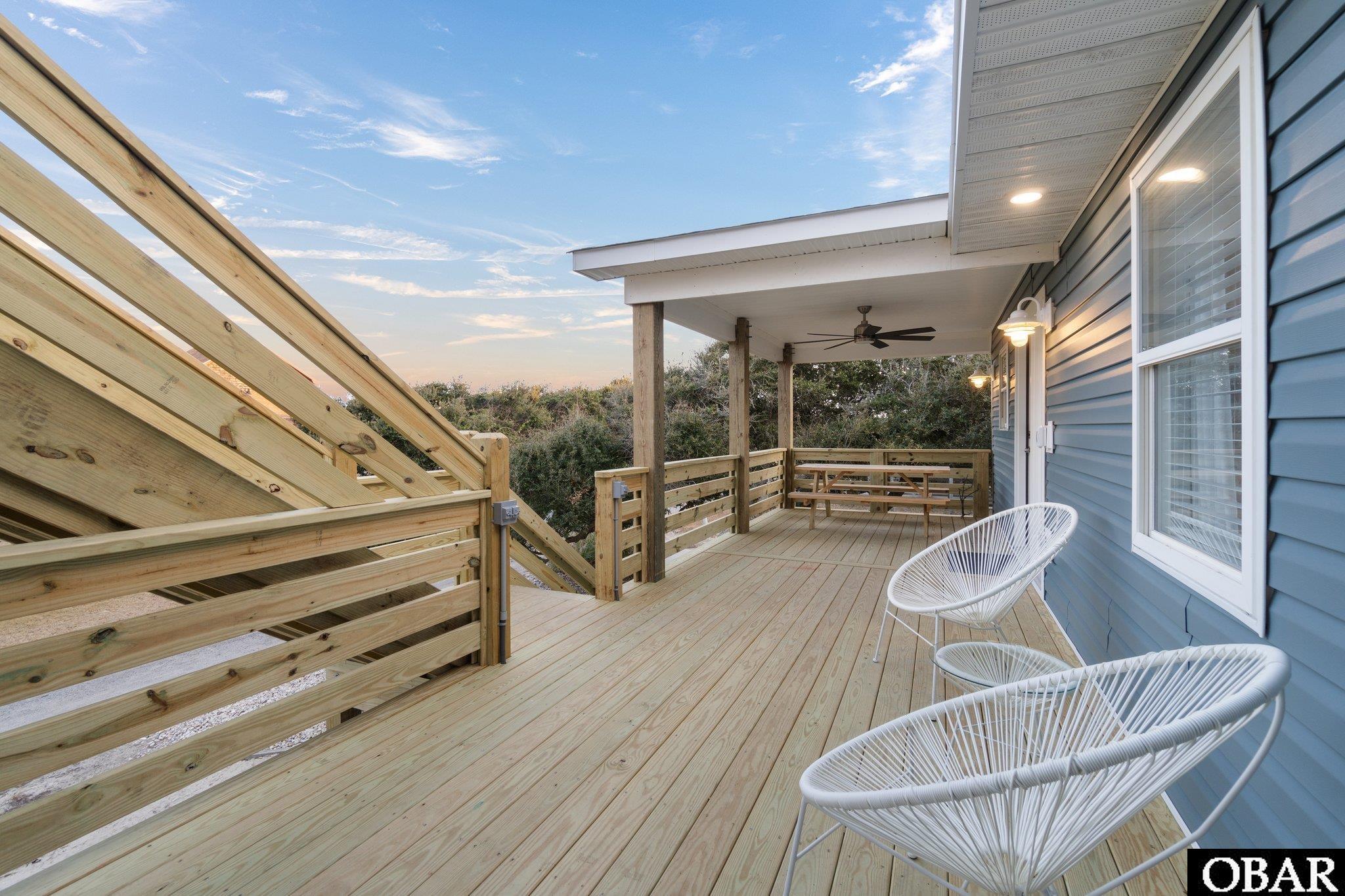 Covered wooden deck with stairs, a picnic table, and white woven chairs beside blue house siding.