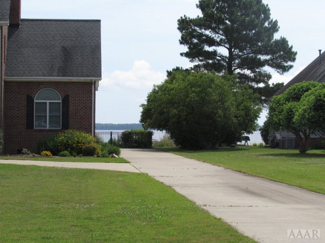 Waterfront property with a concrete driveway, brick house, mature trees, and water in the distance.