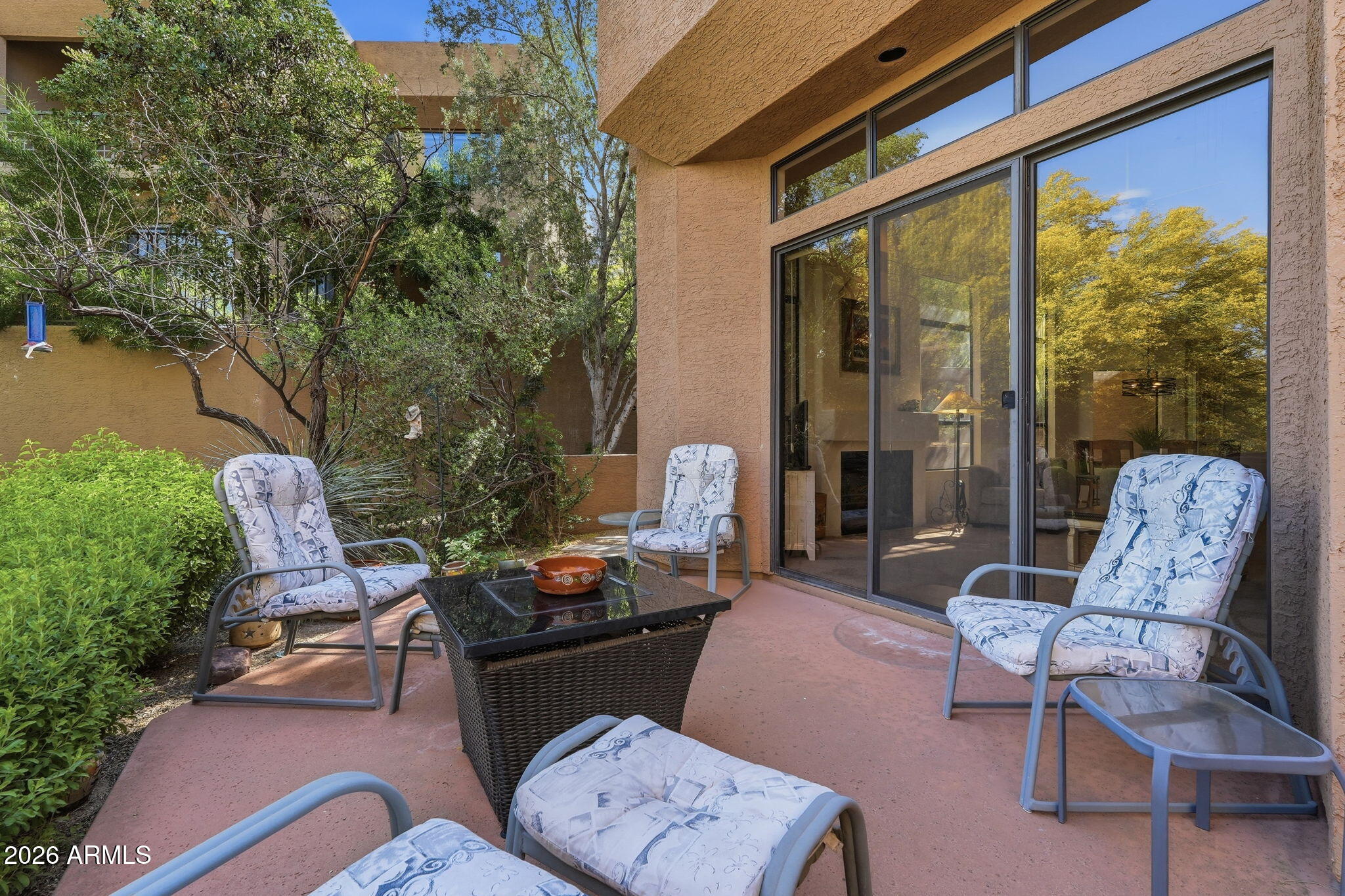 Outdoor patio with cushioned chairs, a wicker coffee table, sliding glass doors, and leafy landscaping.