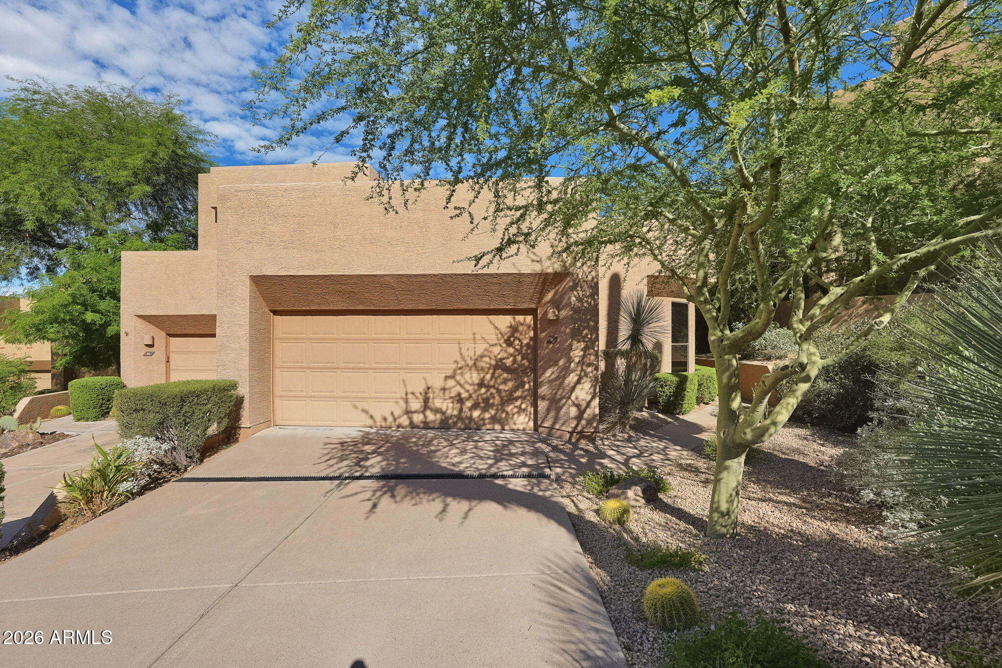 Tan stucco home with a two-car garage, broad driveway, and desert landscaping beneath mature trees.