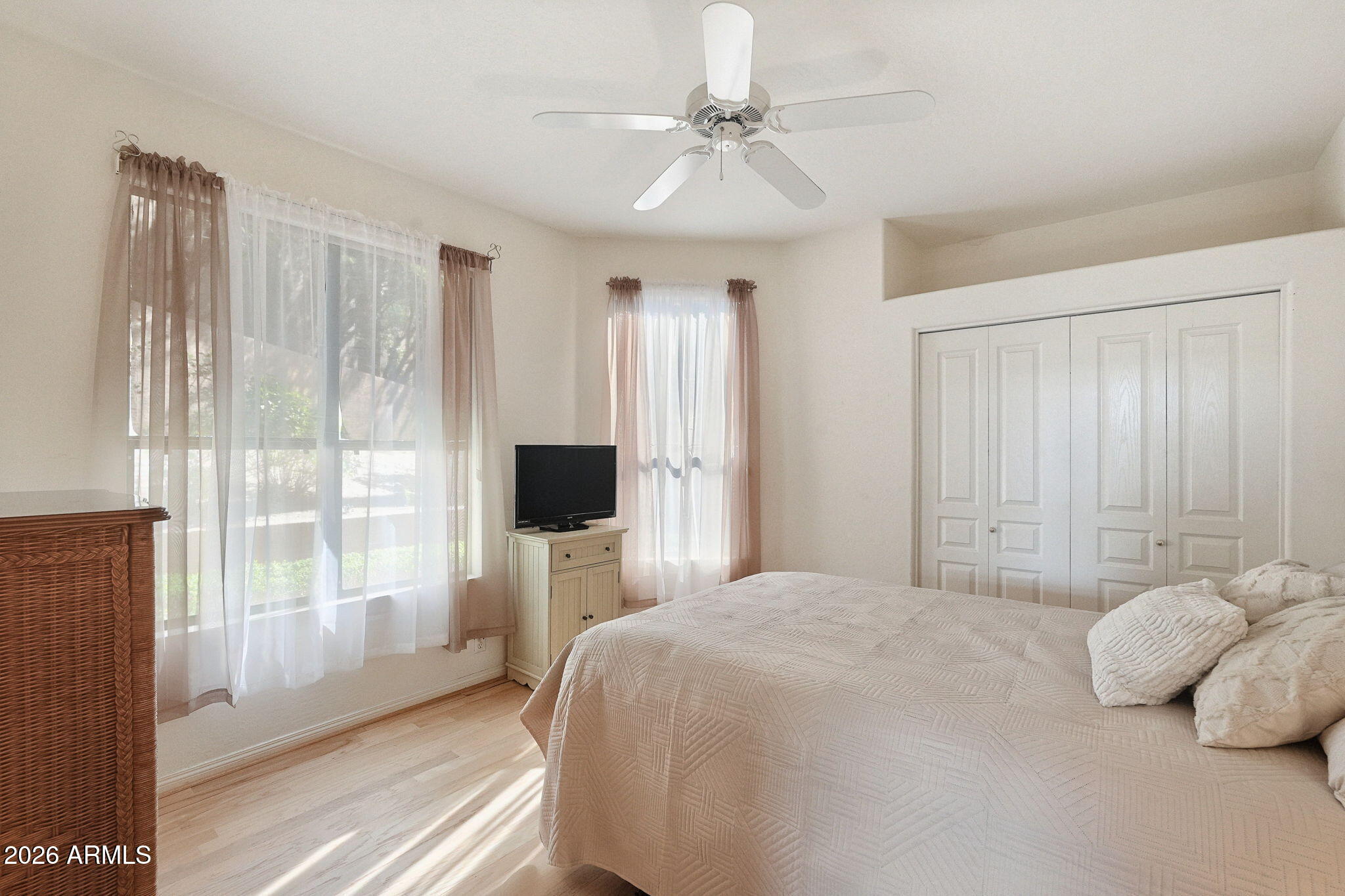 Bedroom with a large bed, sunlit curtained windows, pale wood floors, and a ceiling fan.