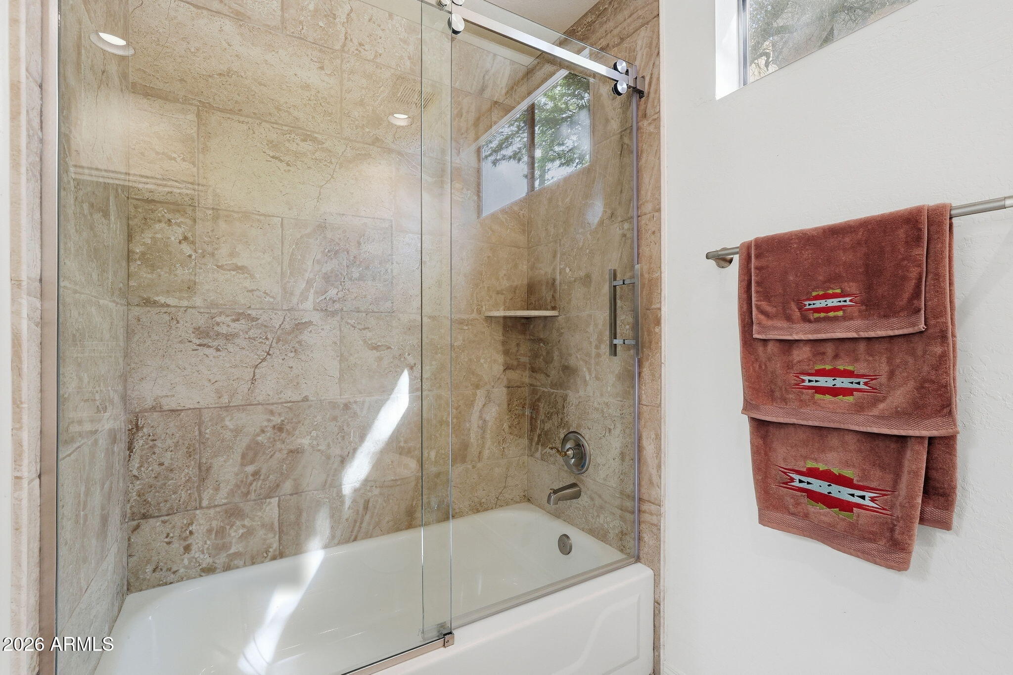 Bathroom bathtub and glass-enclosed shower with beige stone tile and patterned rust-colored towels.