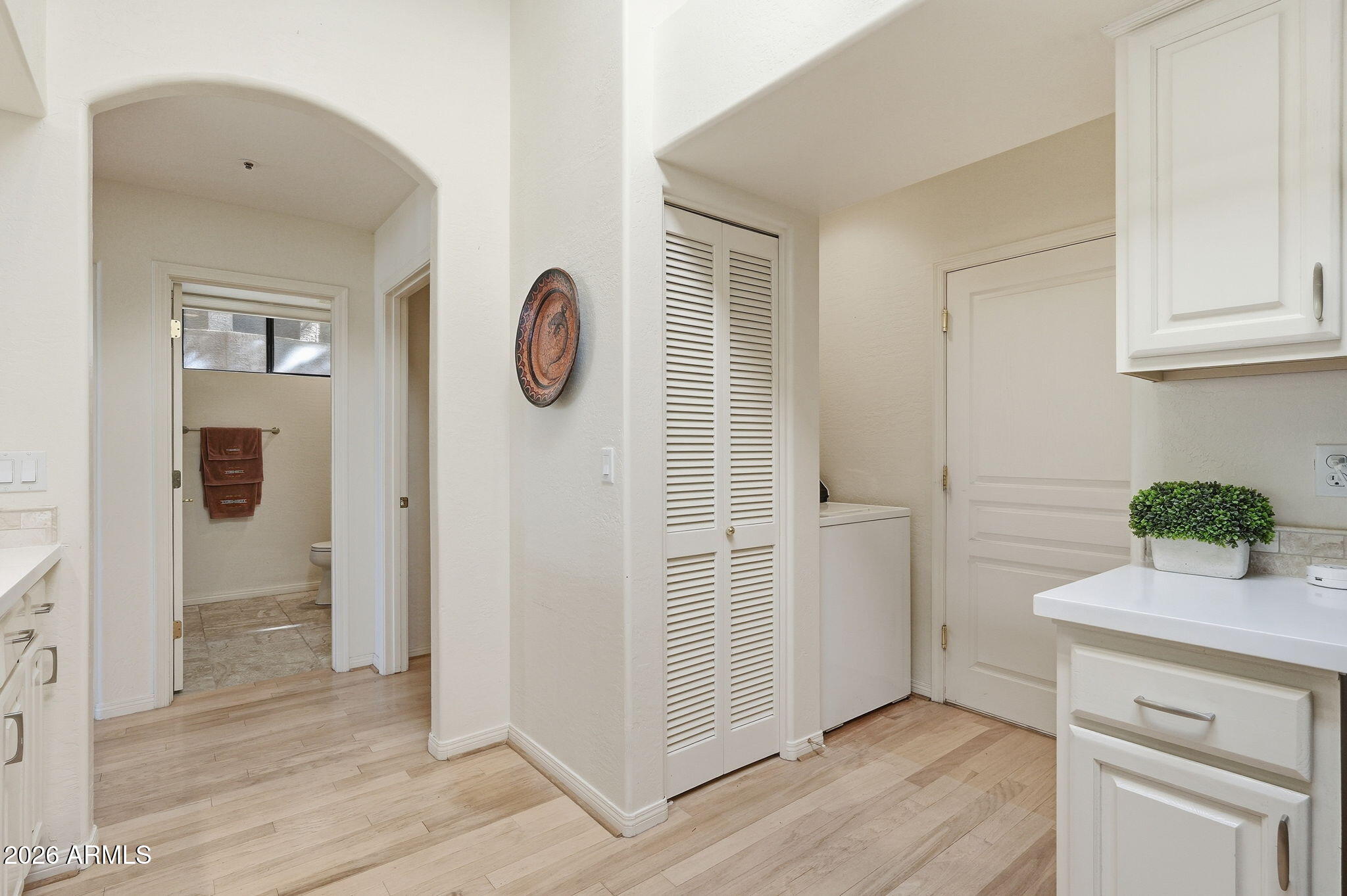 Laundry area with white cabinetry and washer, opening to a bathroom through an arched doorway.
