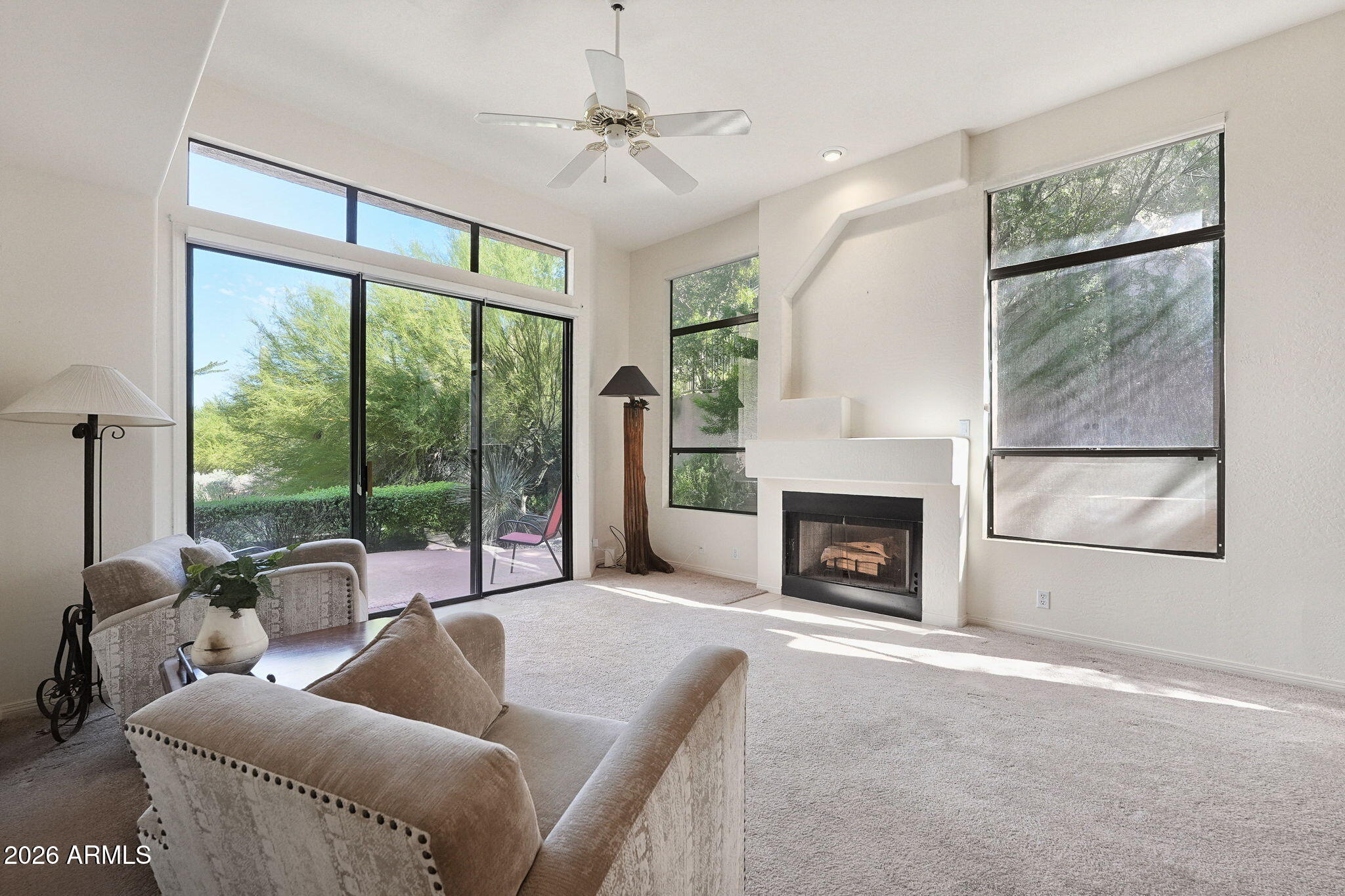 Living room with a white fireplace, tall windows, and sliding glass doors opening to a leafy patio.