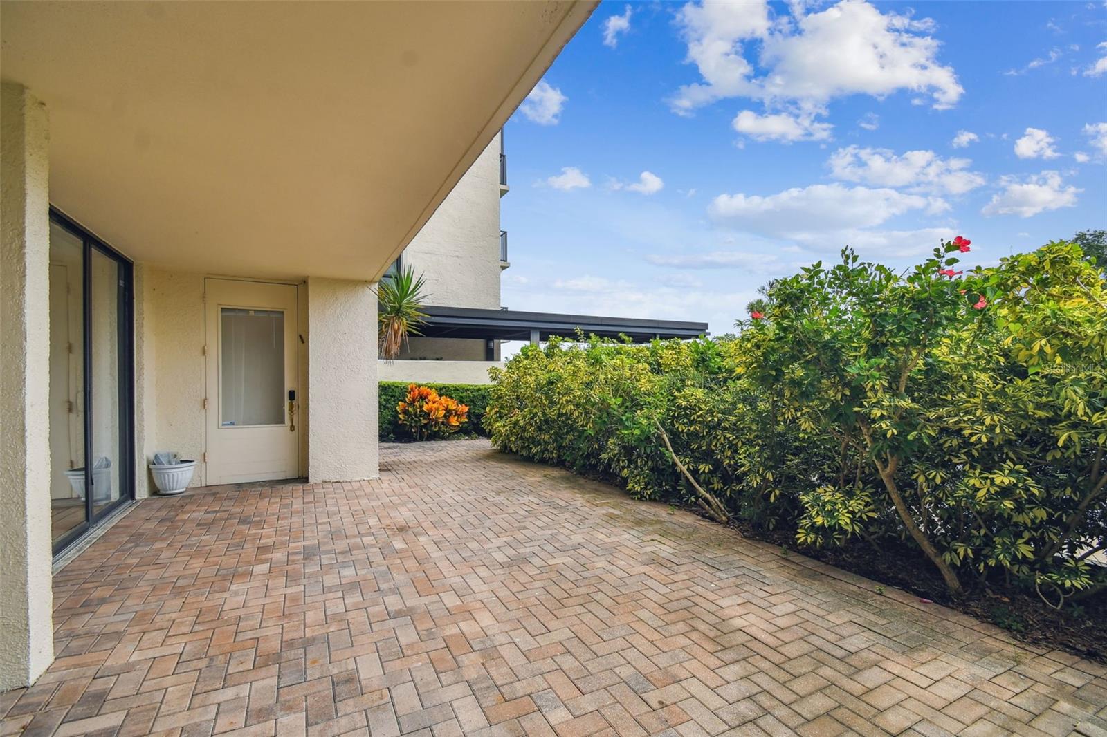 Covered brick patio beside sliding glass doors, bordered by flowering hedges.