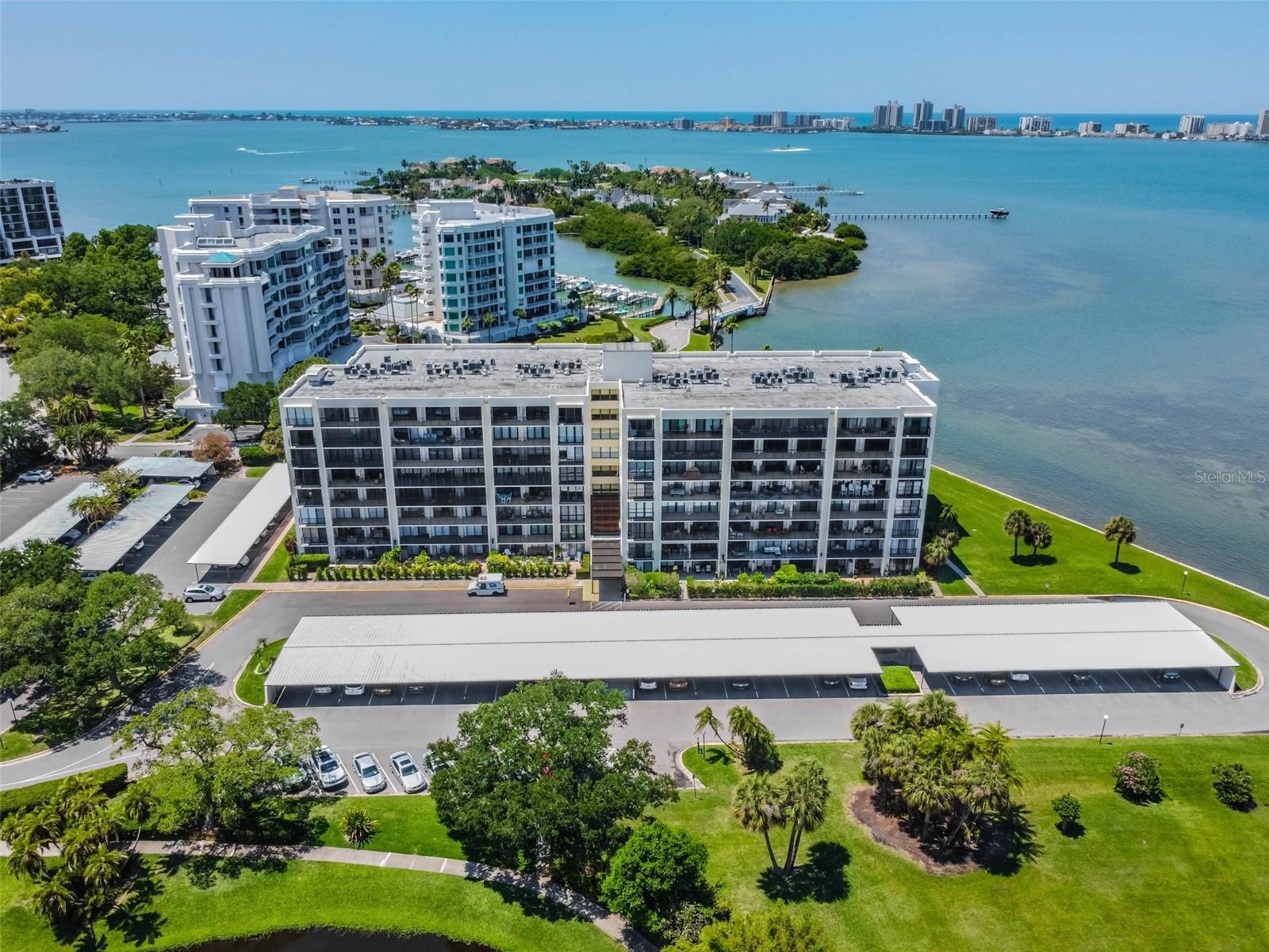 Waterfront condominium building beside a bay, with a long covered parking area and palm-lined grounds.