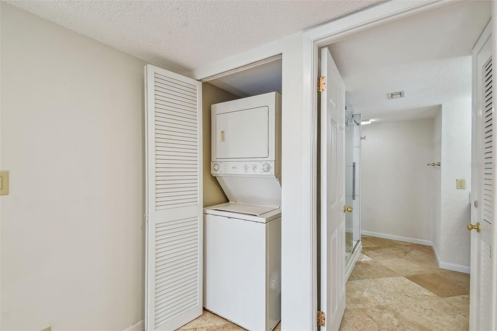Laundry closet with stacked washer and dryer beside a tiled bathroom with a glass shower.