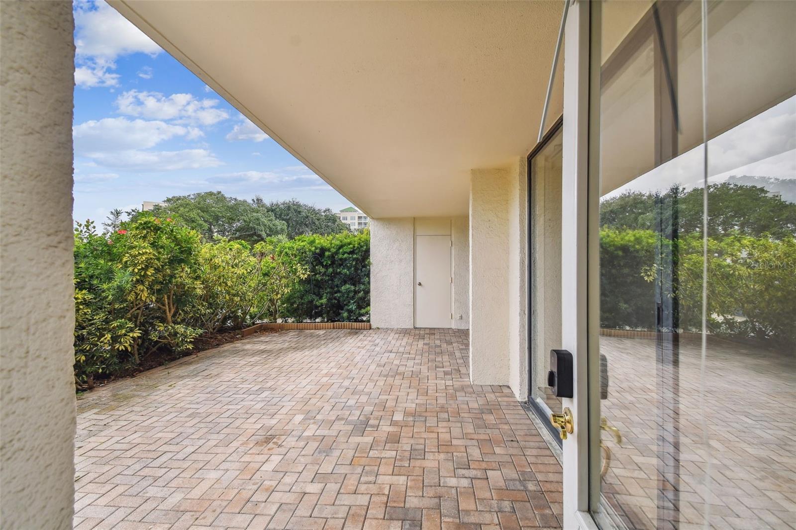 Covered brick-paver patio beside sliding glass doors, with a hedge along the edge.
