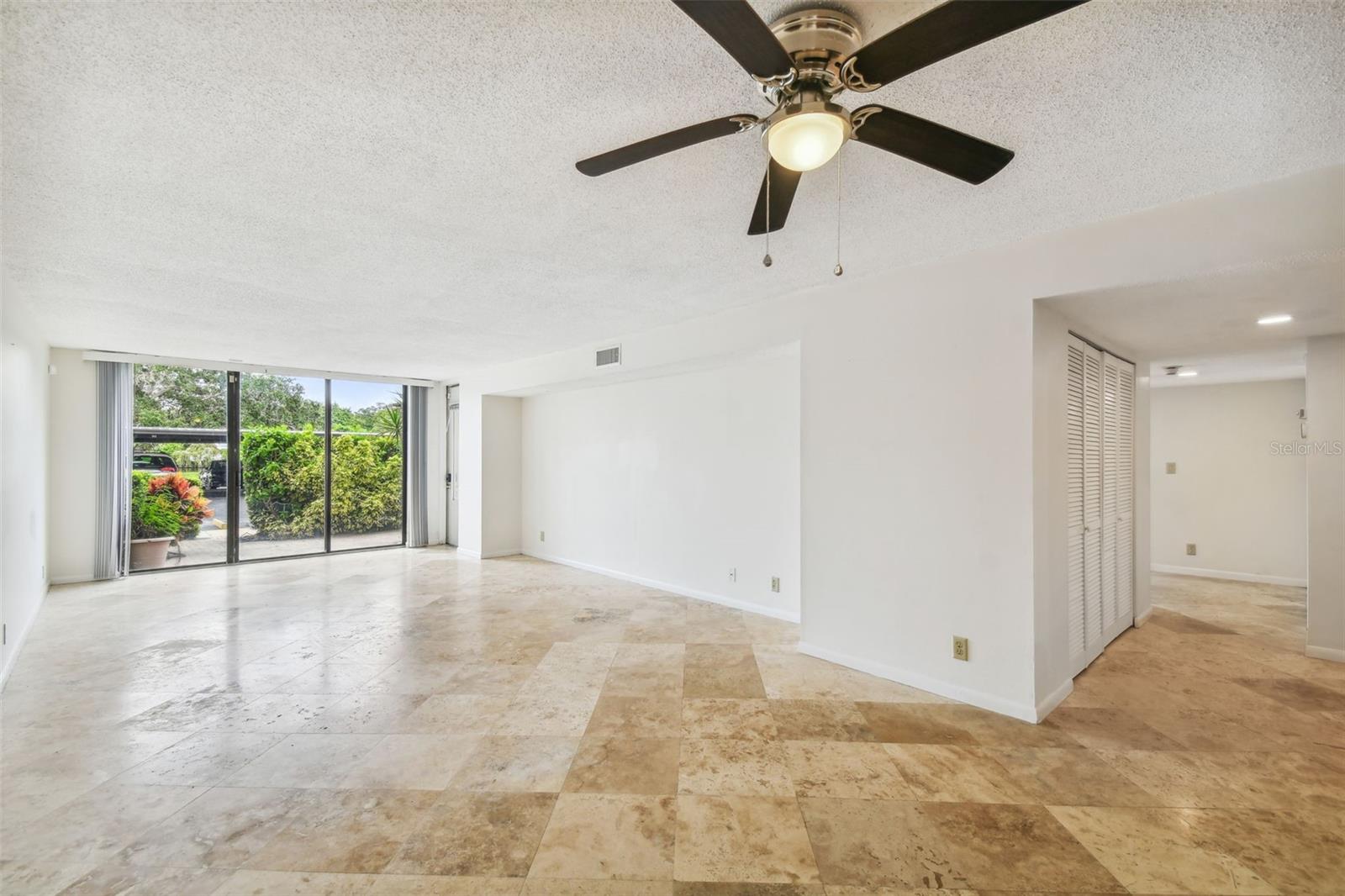 Empty living room with tan stone tile, ceiling fan, and sliding glass doors to a garden.