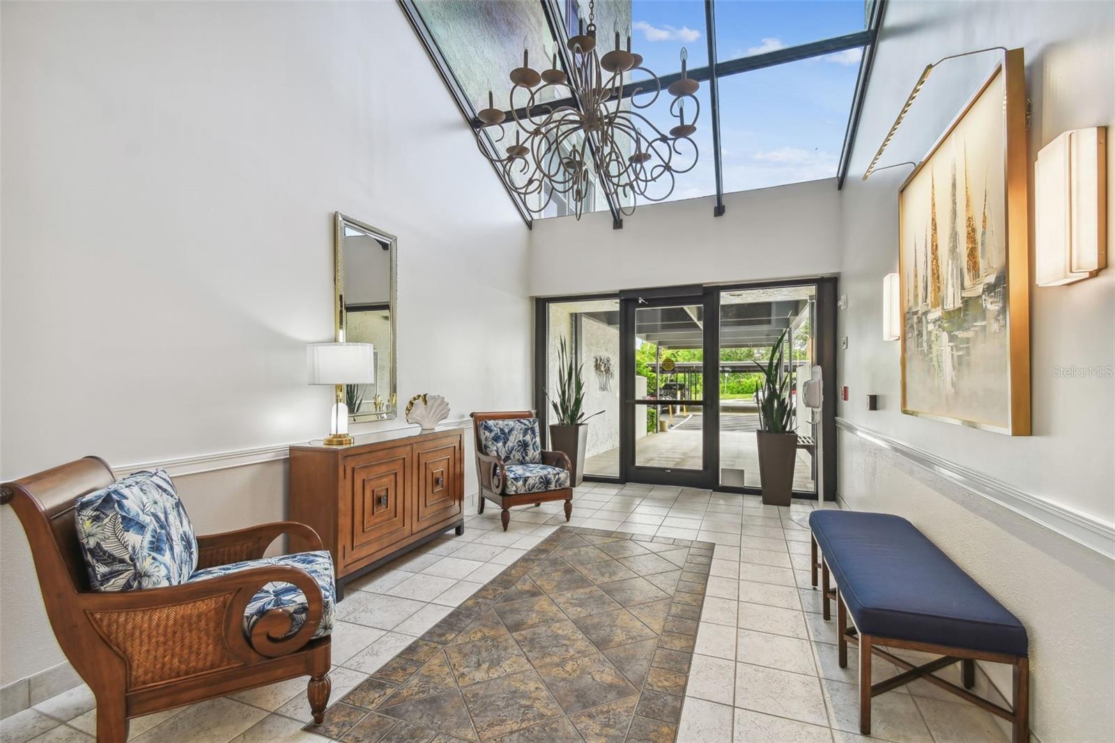 Bright condo lobby with a glass ceiling, ornate chandelier, tiled floor, and seating.