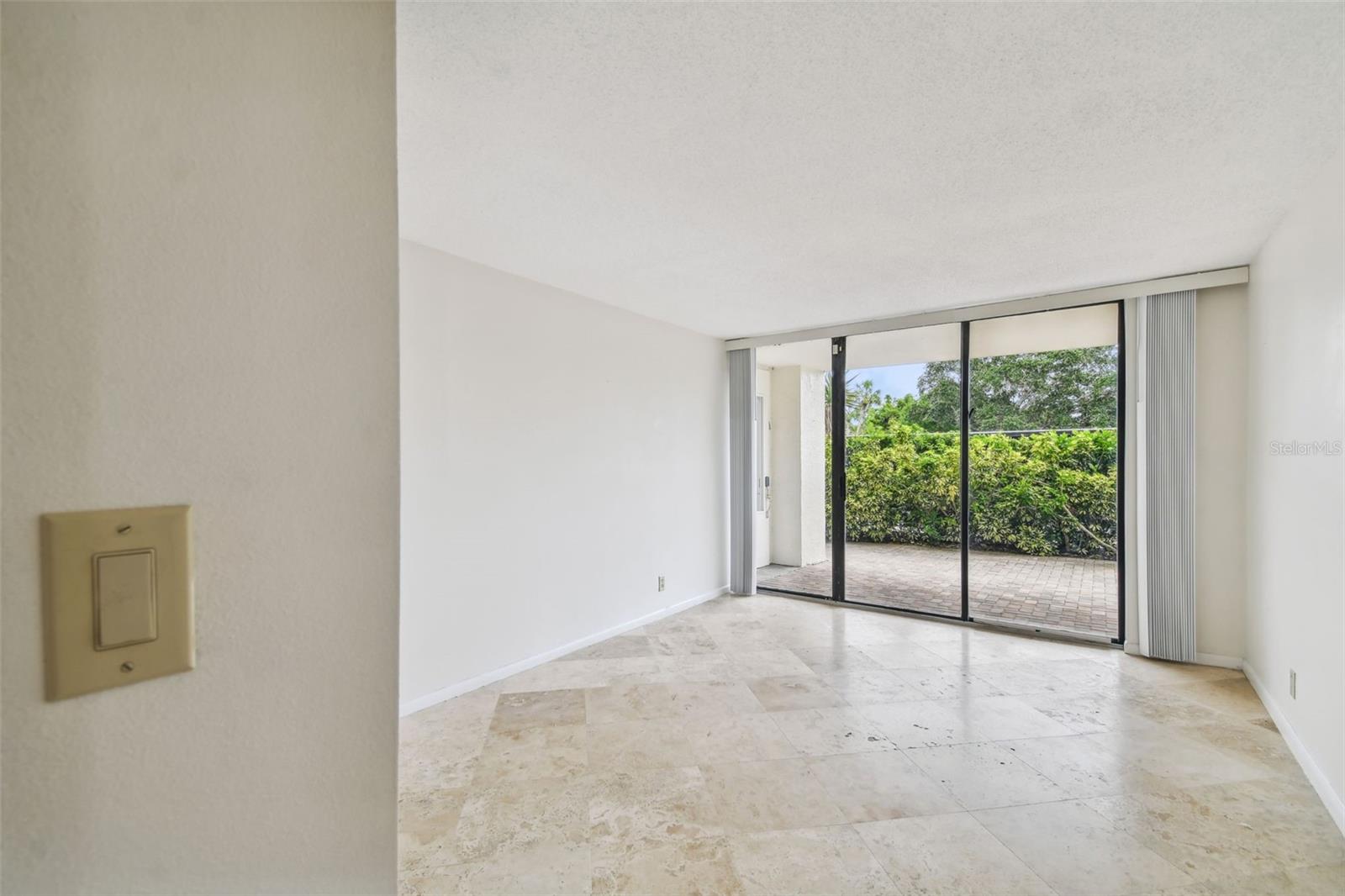 Vacant living room with stone tile flooring and sliding glass doors to a hedge-lined patio.
