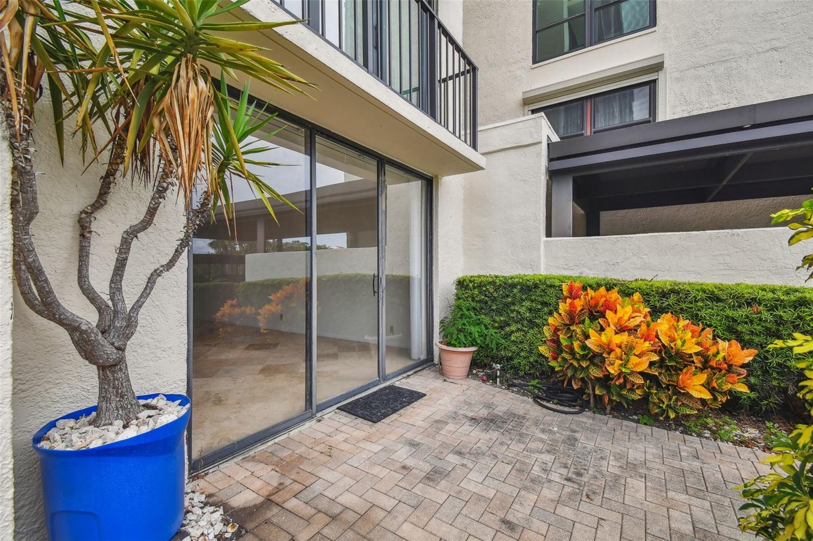 Condo patio with sliding glass doors, brick pavers, tropical plants, and a blue planter.