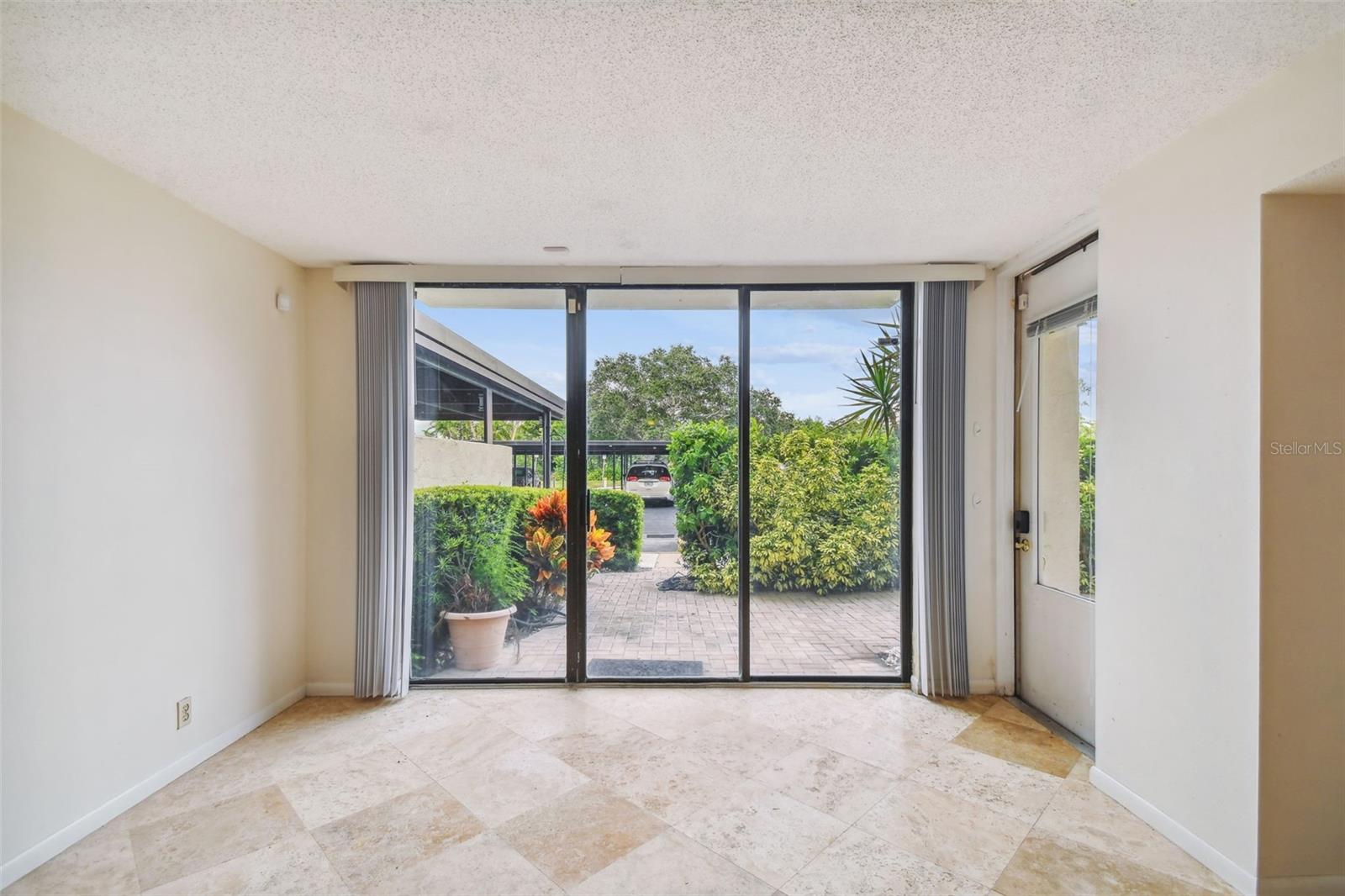 Vacant tiled room with sliding glass doors opening onto a brick patio surrounded by greenery.