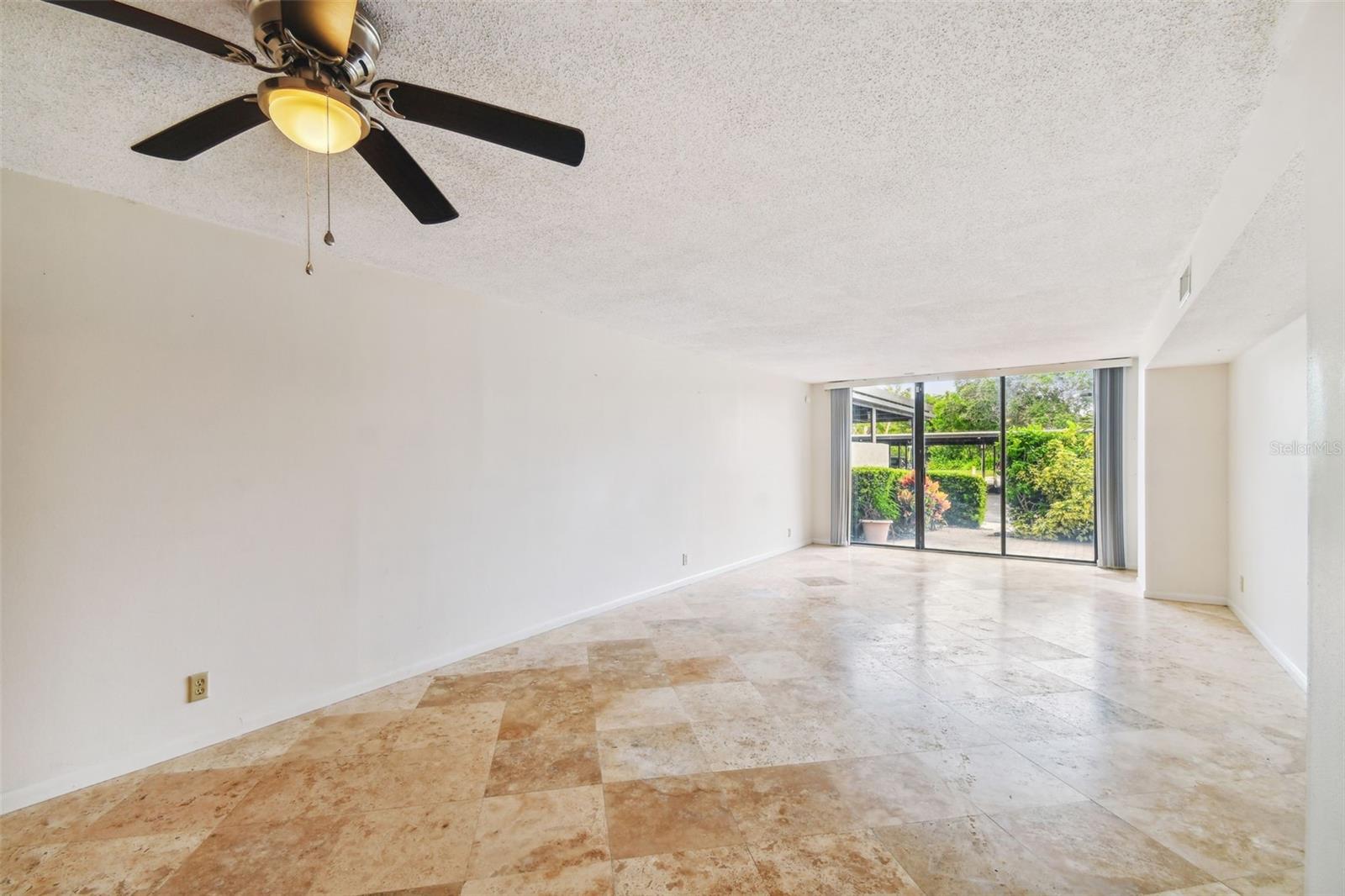 Empty living room with tan tile floors, ceiling fan, and sliding glass doors to a garden patio.
