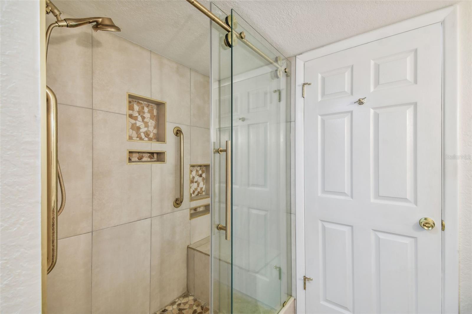 Walk-in shower with beige tile, glass doors, brass fixtures, and mosaic shower niches.