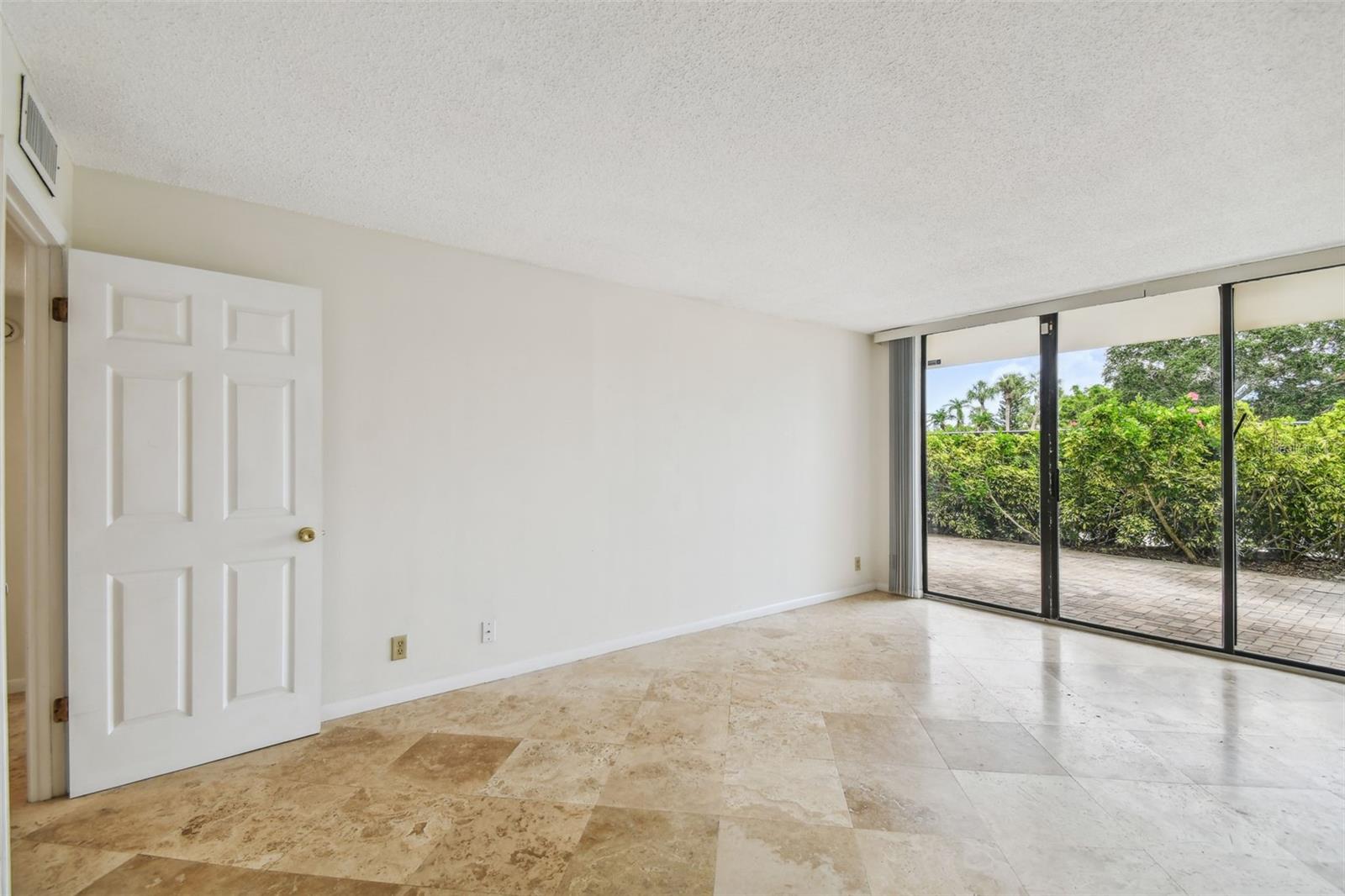 Empty tiled living room with sliding glass doors opening to a hedge-lined patio.