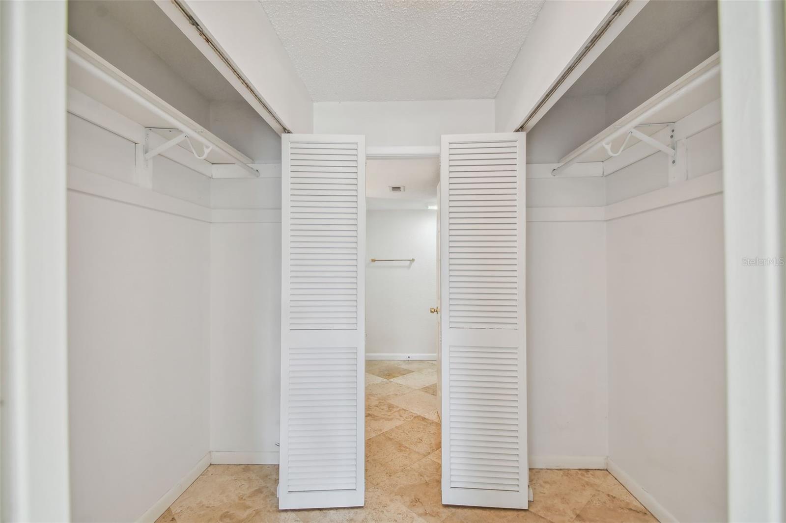 Walk-in closet with white shelving and louvered doors opening to a tiled room.