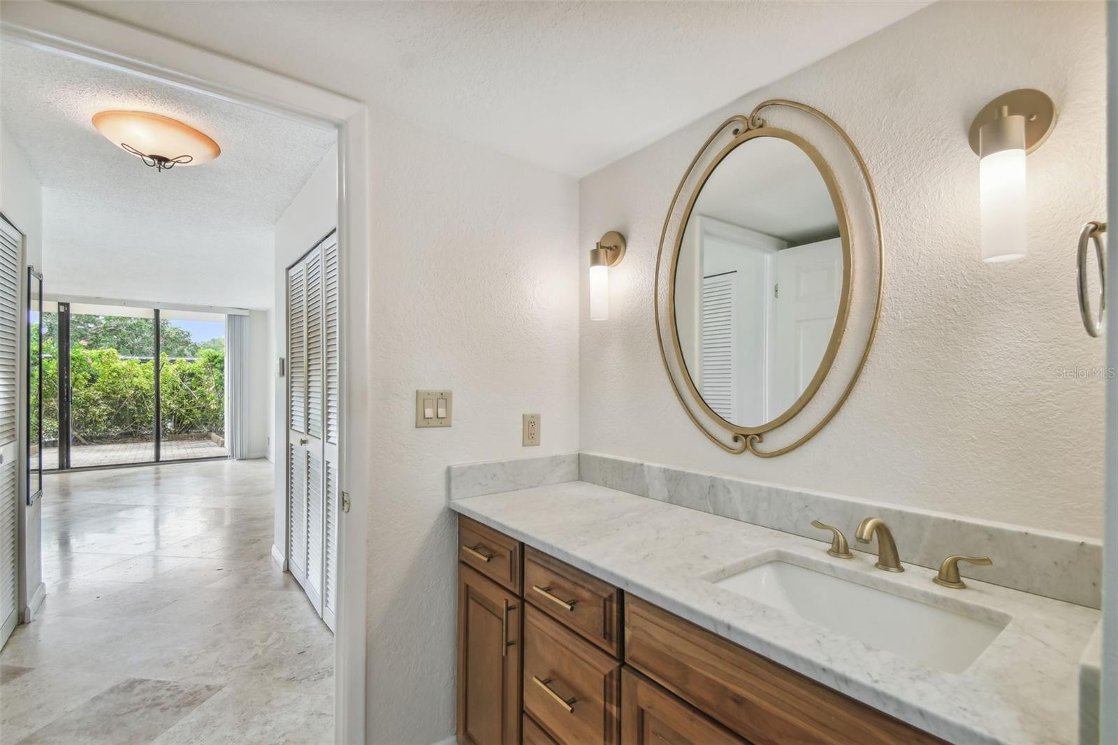 Bathroom vanity with a marble countertop, oval mirror, and wooden cabinets beside a patio-facing room.