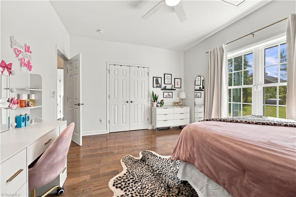 Bright bedroom with a pink bedspread, white furniture, hardwood floors, and large windows overlooking a lawn.