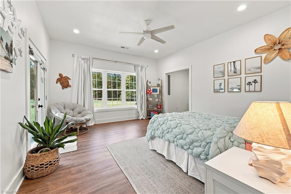 Furnished bedroom with a pale quilted bed, large window, round lounge chair, and wood-look flooring.
