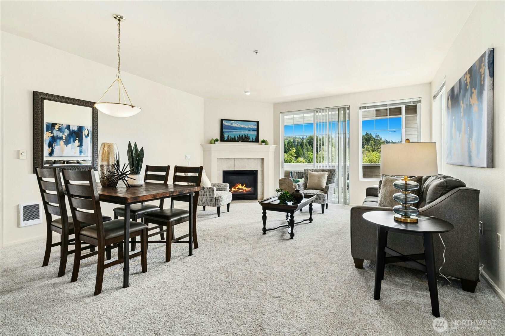 Living and dining area with a fireplace, gray seating, and large windows overlooking trees.