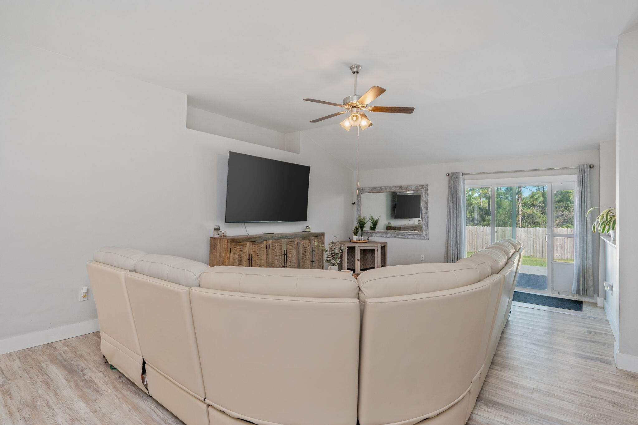 Living room with a curved cream sectional, wall-mounted TV, ceiling fan, and sliding glass doors to the backyard.