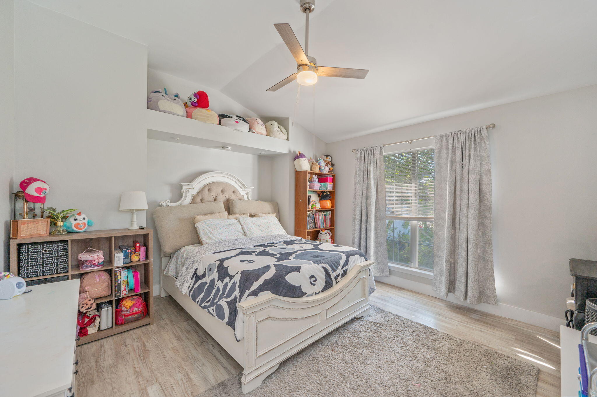 Bedroom with a white bed, toy-filled shelves, and a large curtained window.